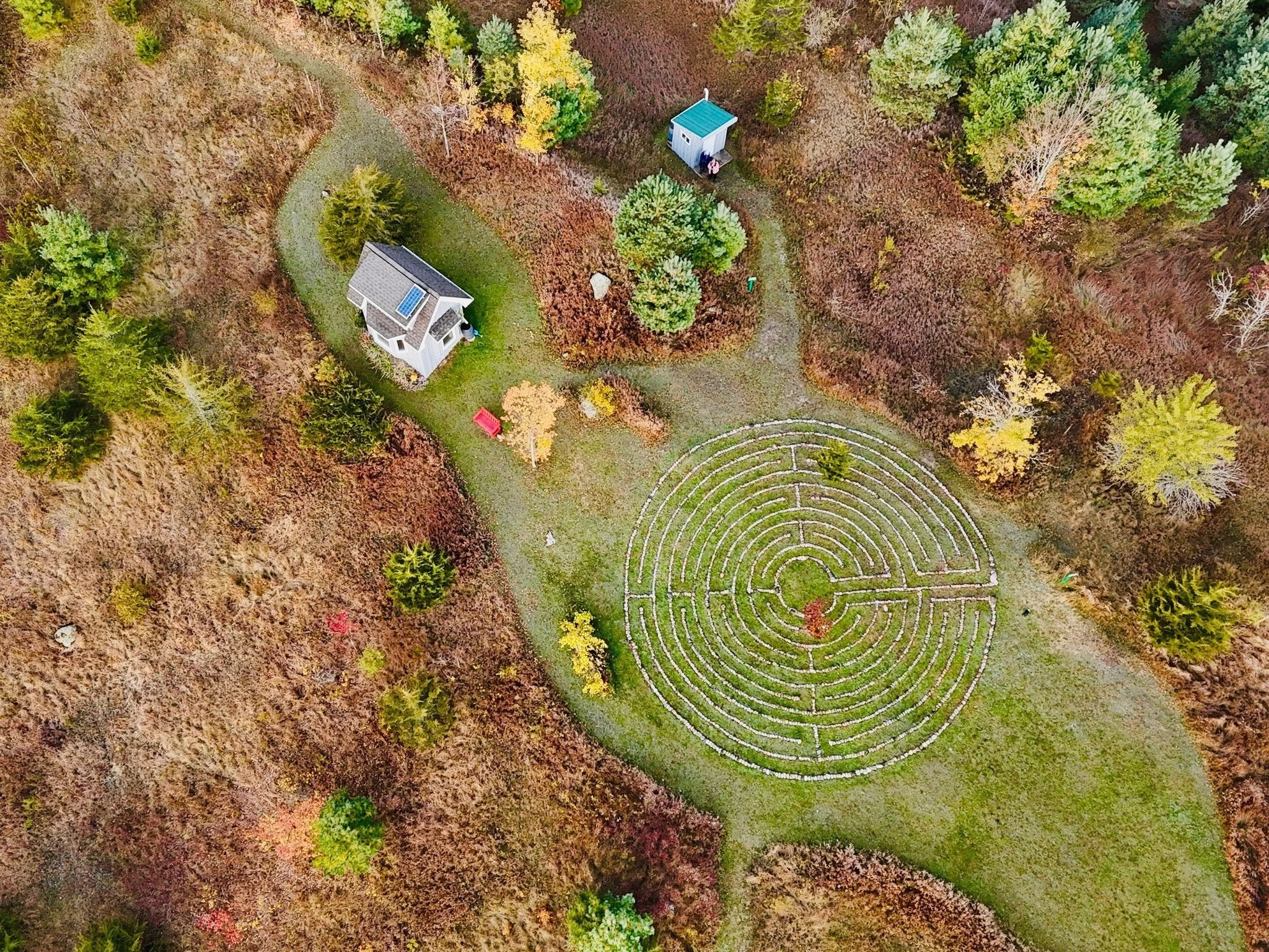 An aerial view of a park with a circular hedge maze, a small house with a solar panel on the roof, and a small blue shed, surrounded by trees with fall foliage.