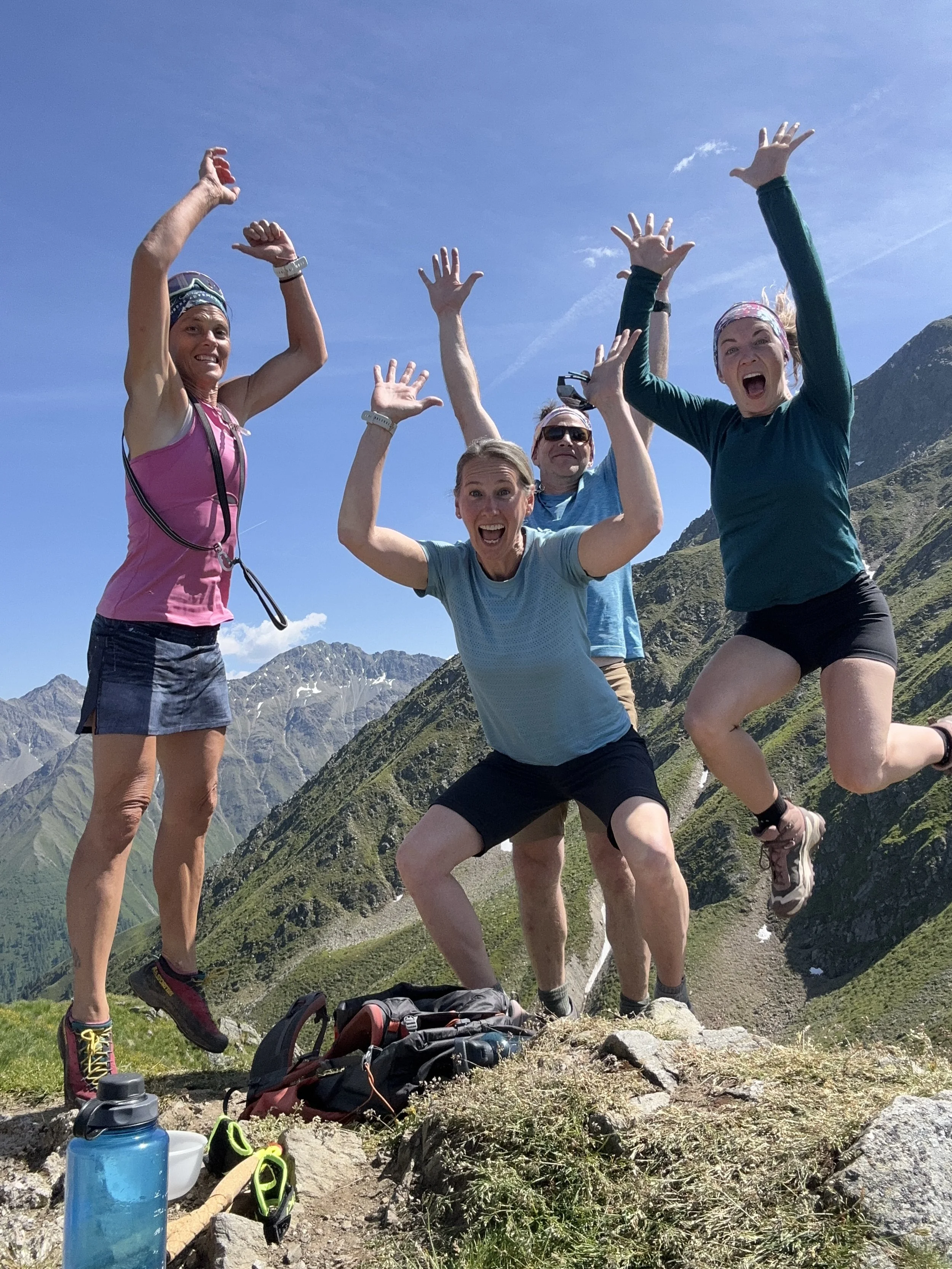 Four people, two women and two men, celebrating and jumping on a mountain trail with a mountainous landscape and clear blue sky in the background.