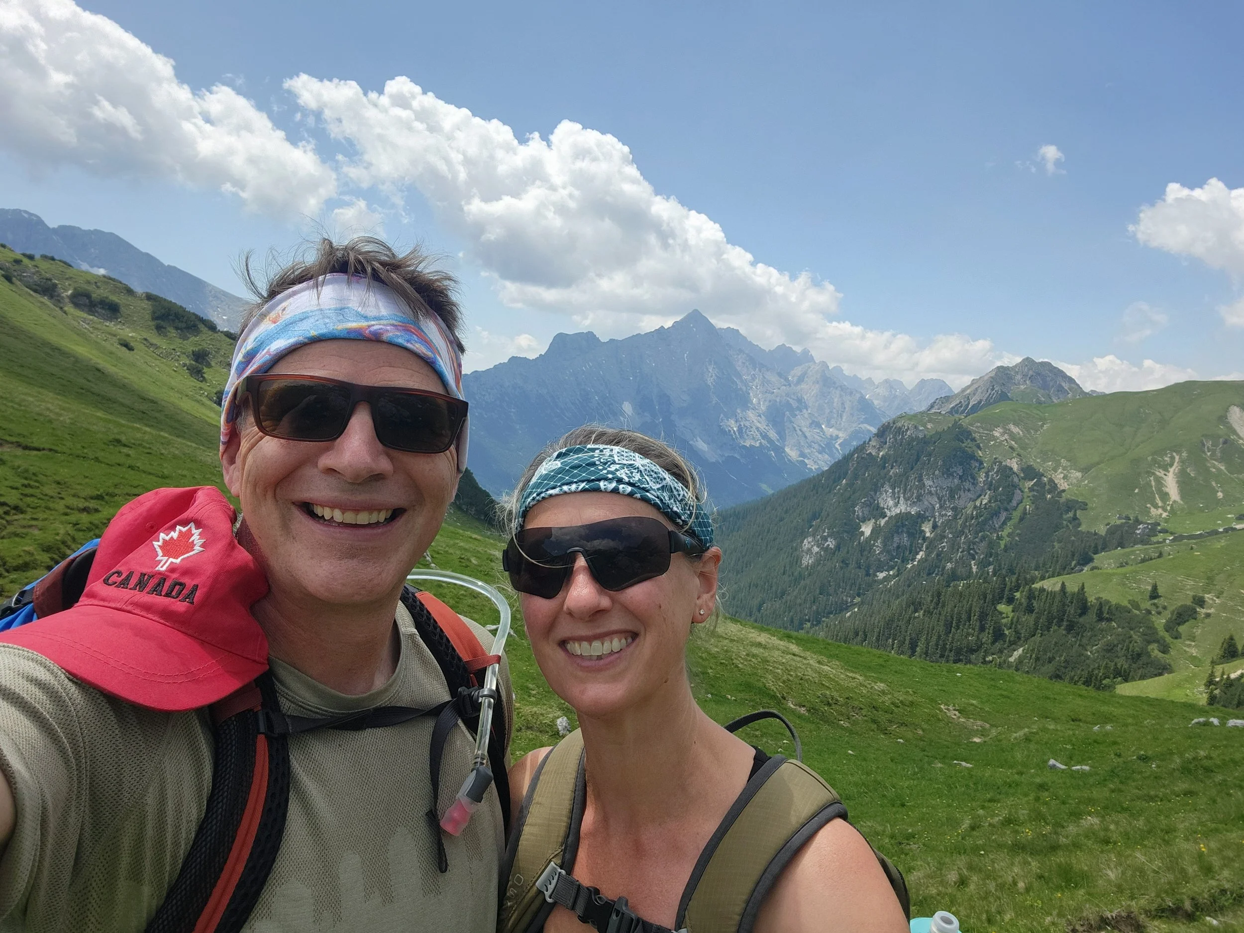 A smiling man and woman taking a selfie during a hike in the mountains on a clear day, both wearing sunglasses and bandanas.