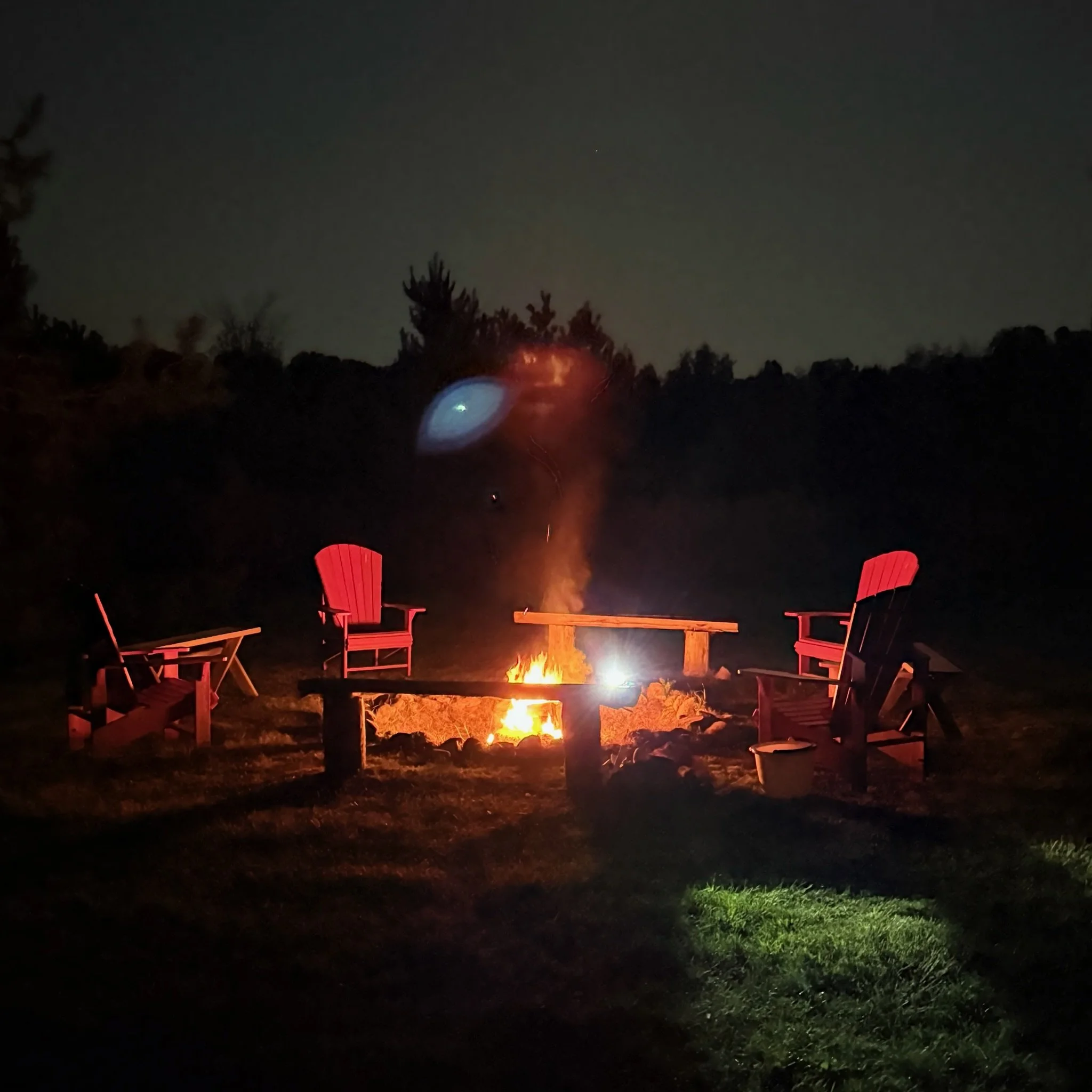 Nighttime outdoor fire pit with four red Adirondack chairs arranged around it on a grassy area, trees in the background.