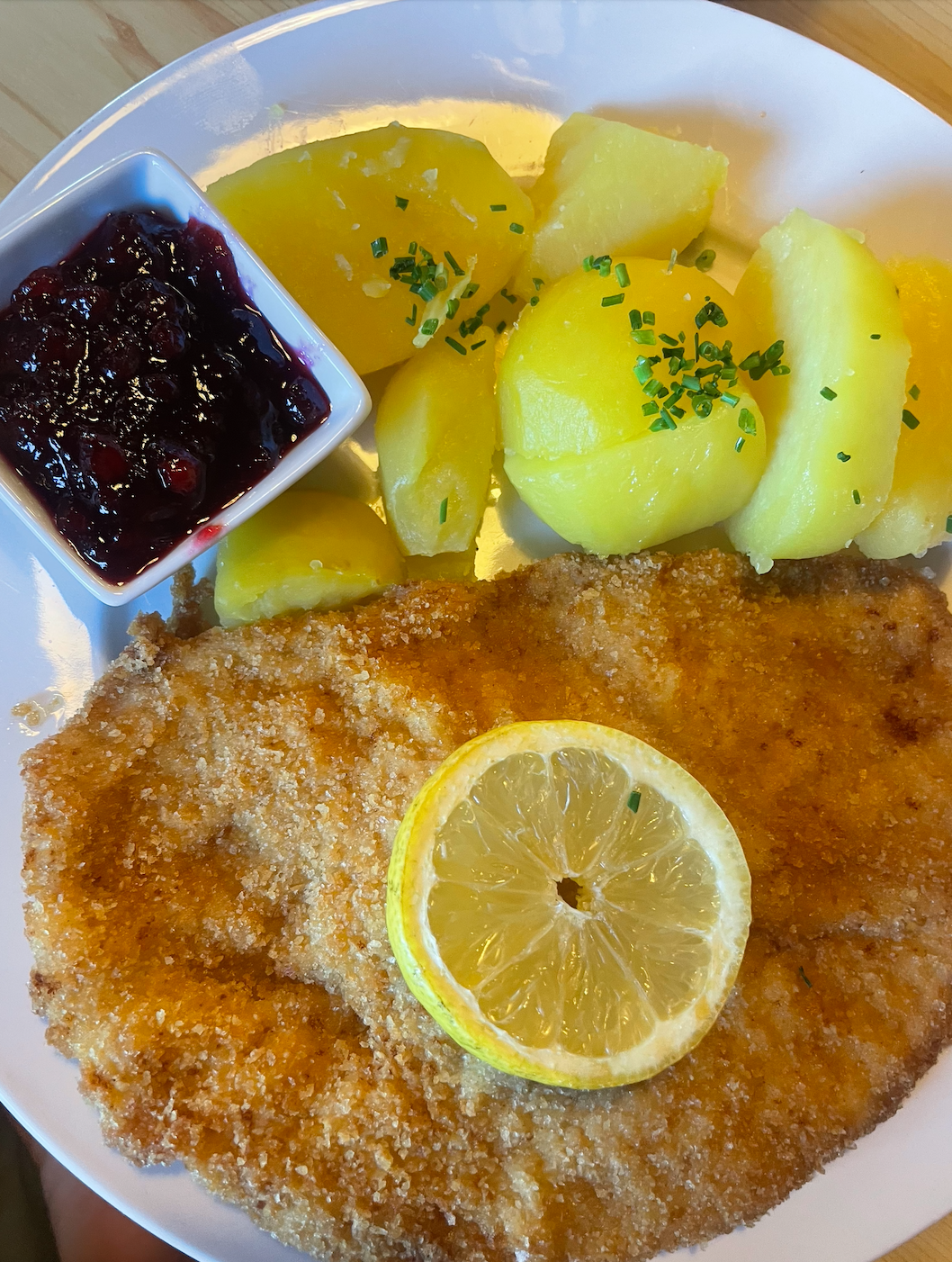 Plate of fried fish fillet garnished with lemon, with boiled potatoes topped with chopped chives, a small dish of berry sauce, and a wooden table in the background.