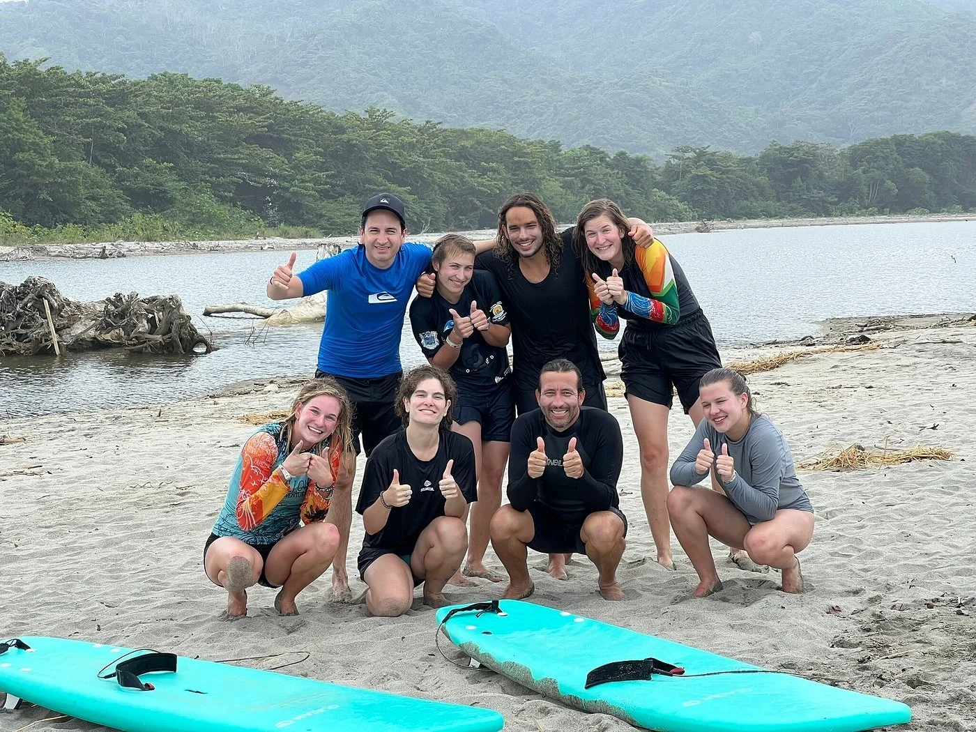 Group of nine people posing on a sandy beach with two surfboards, smiling and giving thumbs up in front of a lake with trees and mountains in the background.