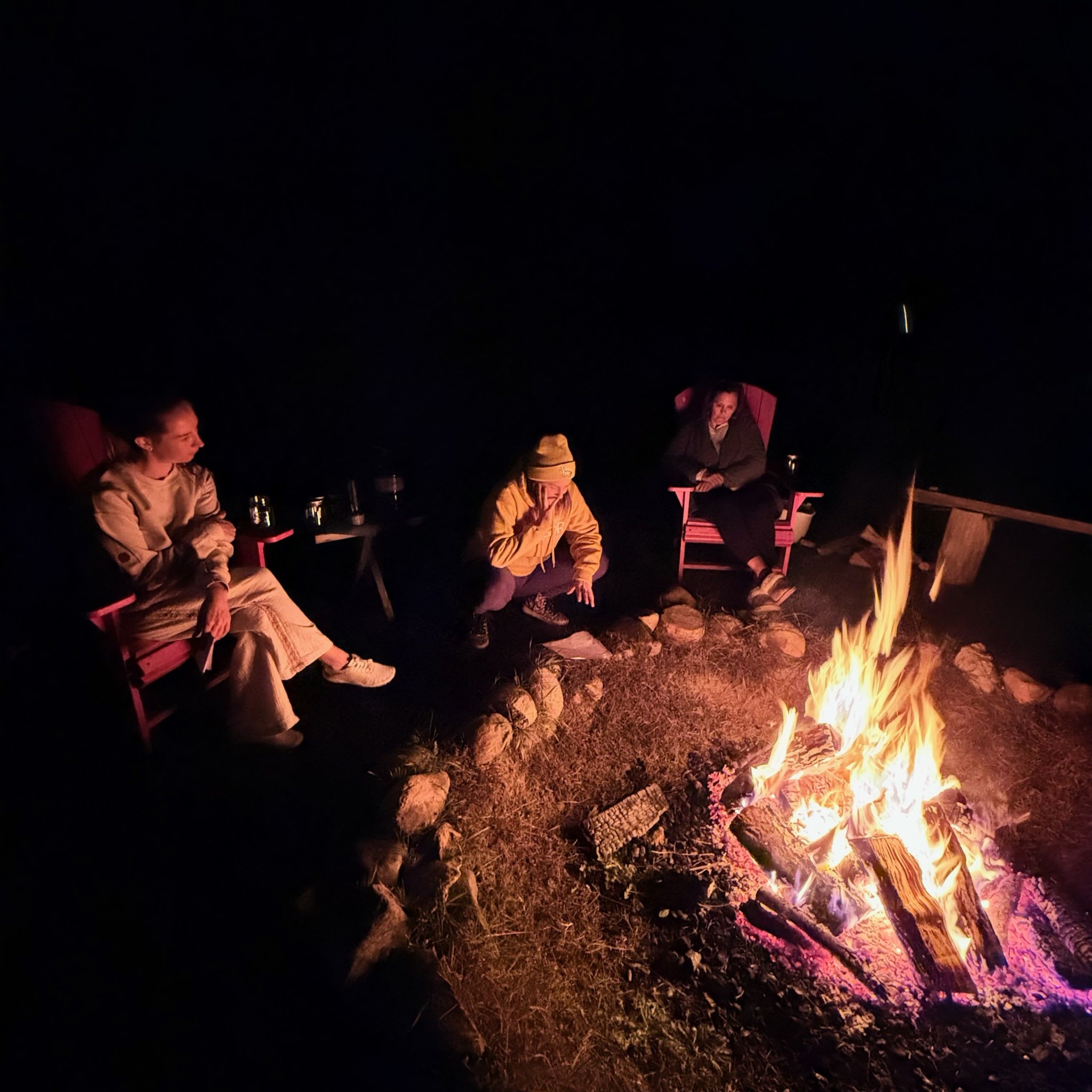 Three people sitting around a campfire at night, two women in chairs and a man crouching, with drinks on a small table nearby.