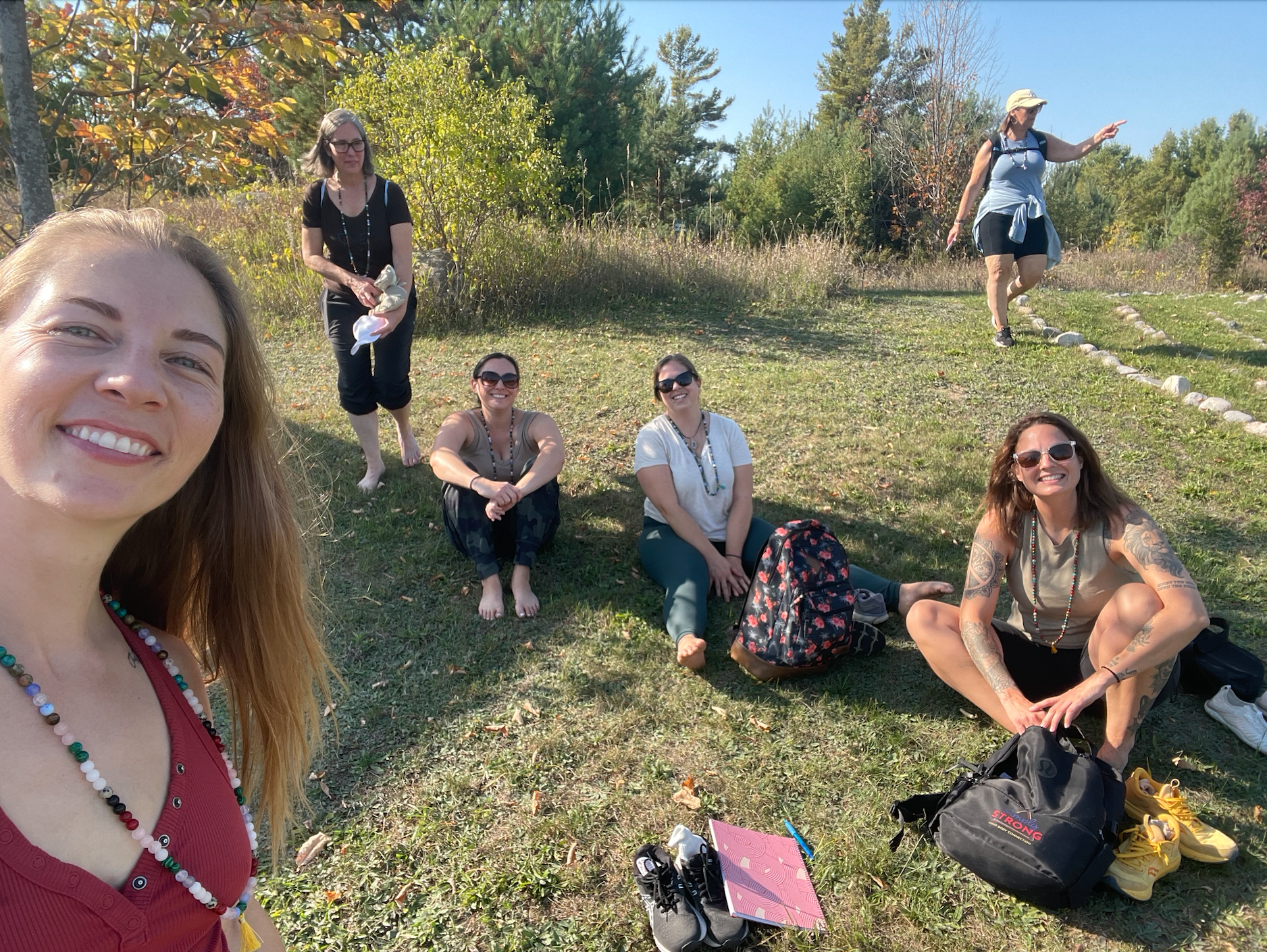 Group of women enjoying a sunny outdoor day in a grassy park, with some sitting and others standing, surrounded by trees with autumn leaves.