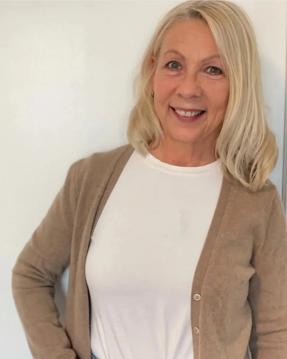 A smiling woman with blonde hair wearing a white shirt and a beige cardigan, standing against a plain white background.