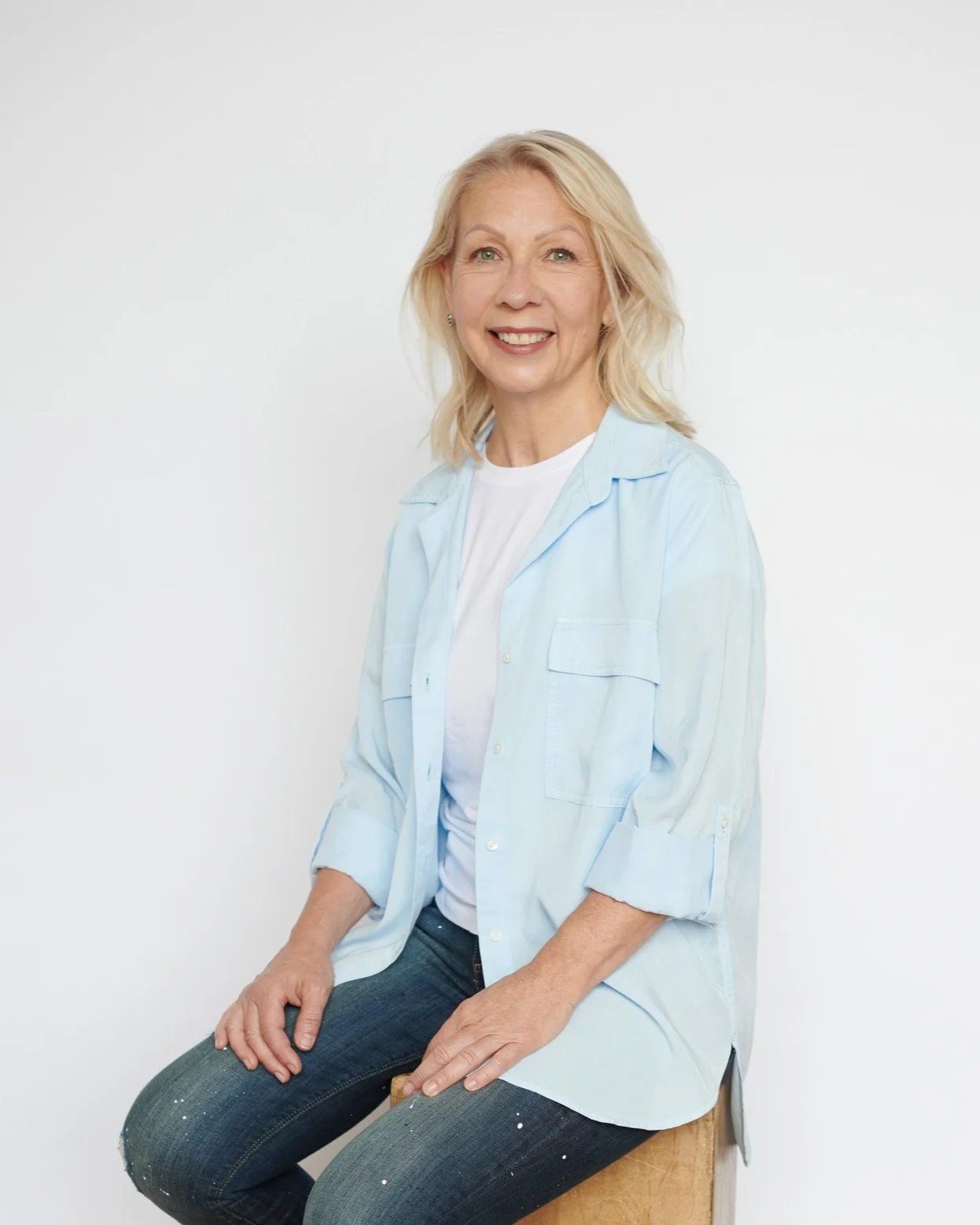 A woman with blonde hair, smiling, sitting on a wooden stool against a plain white background, wearing a light blue button-up shirt over a white T-shirt and dark jeans.