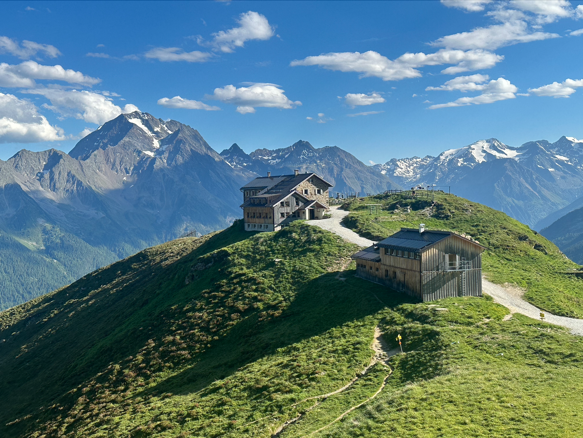 Mountains with snow caps, blue sky with clouds, and two wooden buildings on a grassy hillside with a dirt pathway.