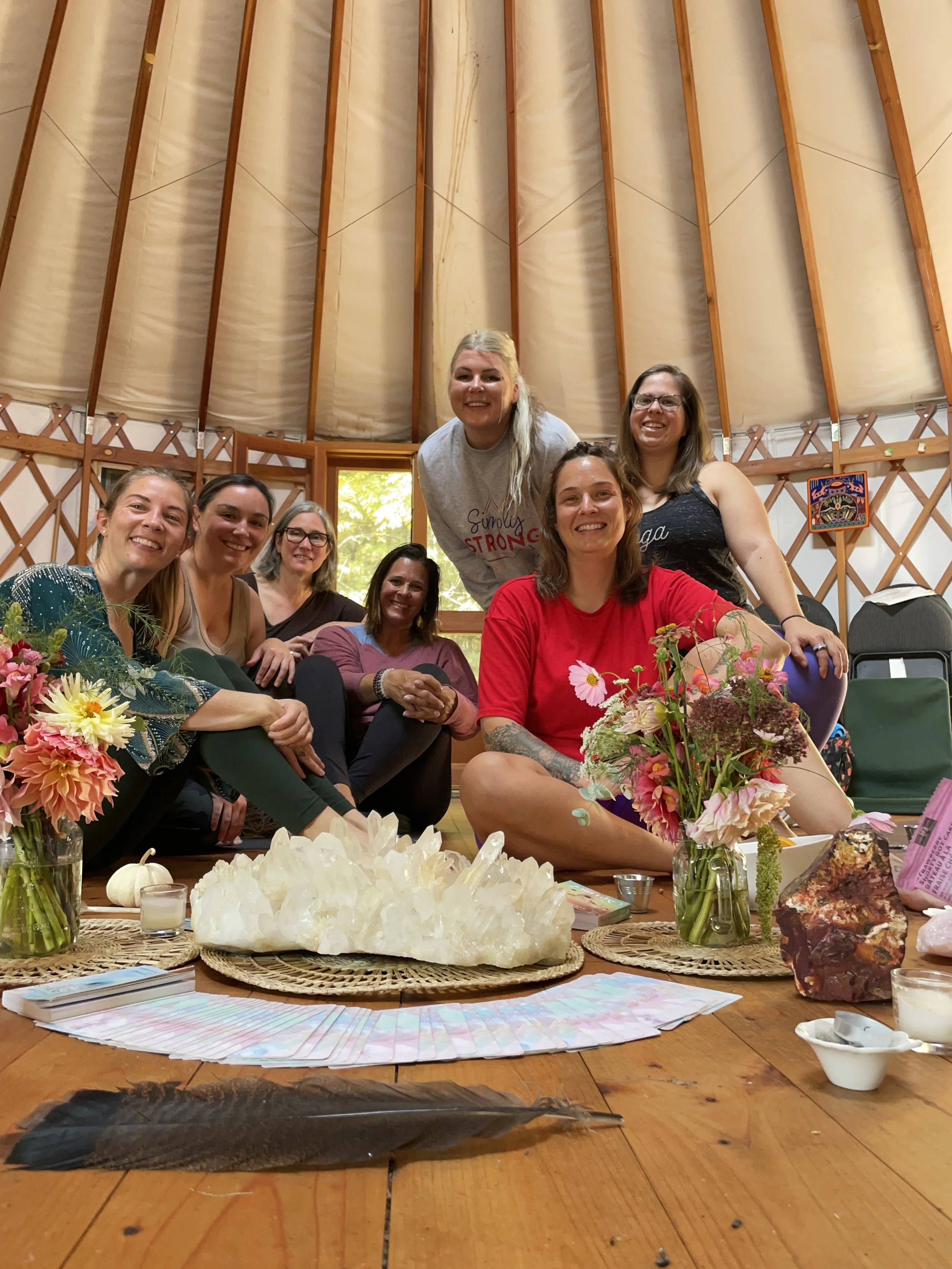 Group of seven women gathered in a circle inside a yurt, smiling and posing for a photo, with flowers, crystals, and candles on the table in front of them.