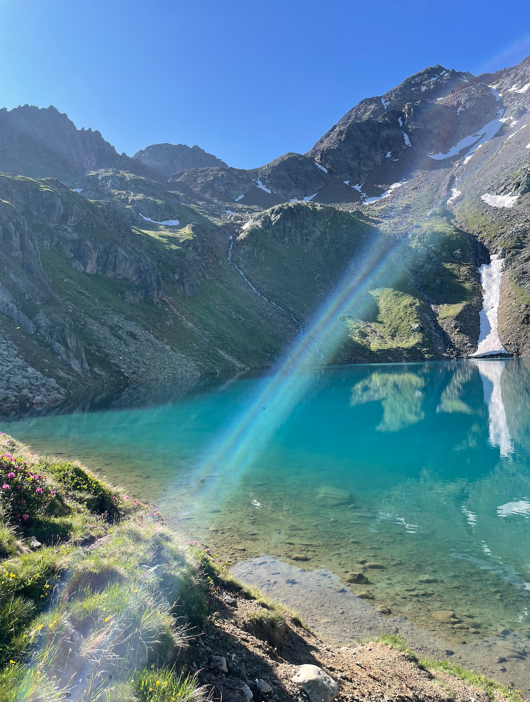 Mountain landscape with snow patches, green slopes, a waterfall, a lake, and a rainbow in clear blue sky.