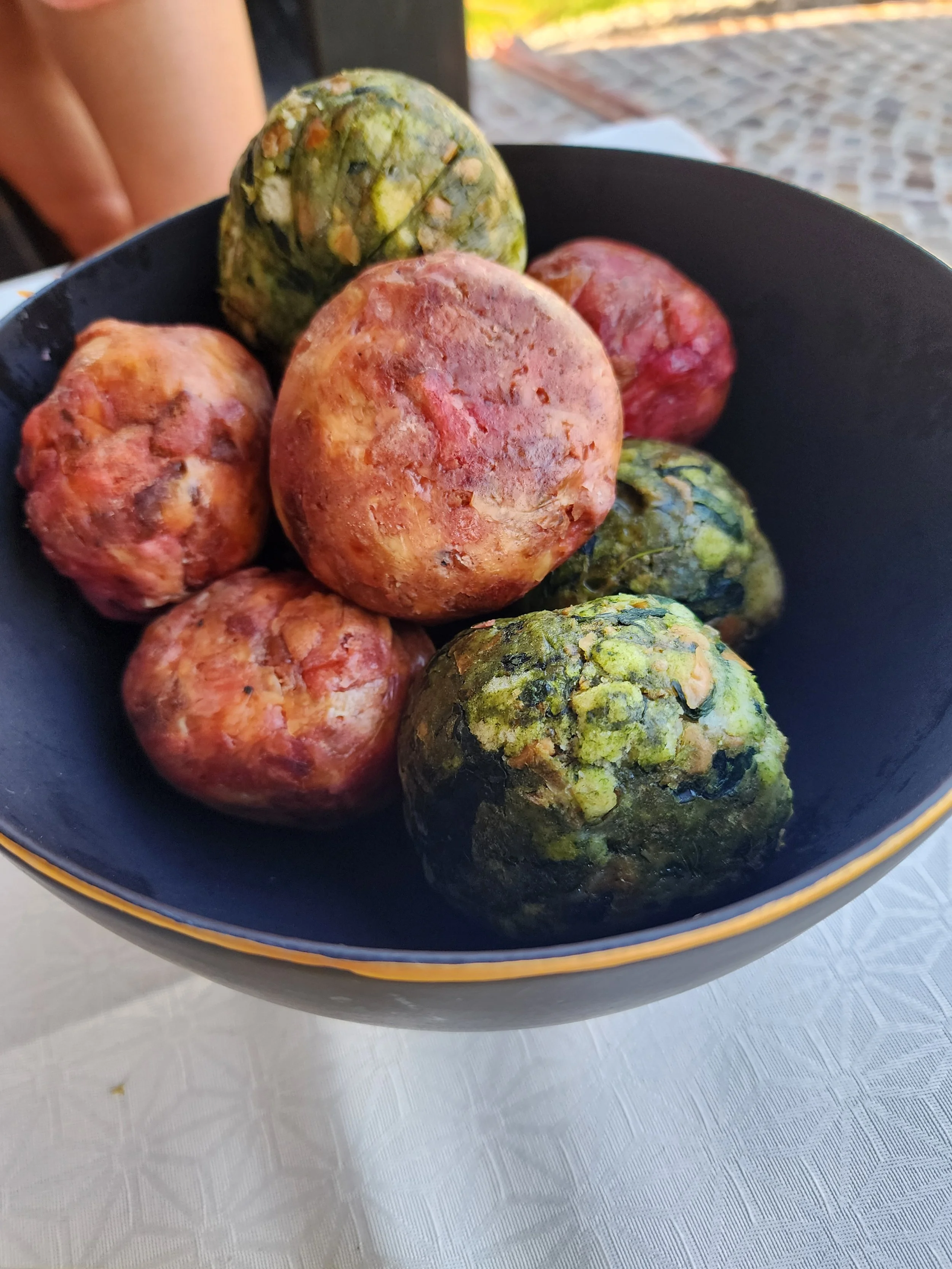 A black bowl with a gold rim filled with assorted colorful round root vegetables, including purple, red, and green-topped tubers, on a white tablecloth