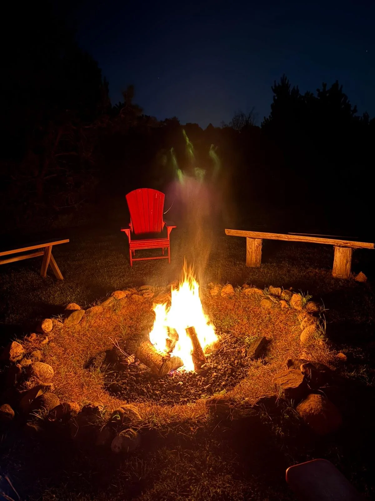 Nighttime outdoor scene with a campfire surrounded by rocks, with a red Adirondack chair and wooden benches nearby, and a dark forest in the background.