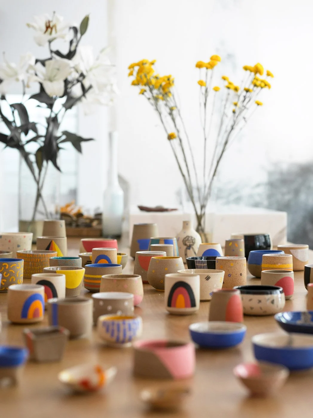 Multiple ceramic pieces by LODAZAL Ceramica with colorful designs. Placed on a large wooden table with large flower arrangements in the background