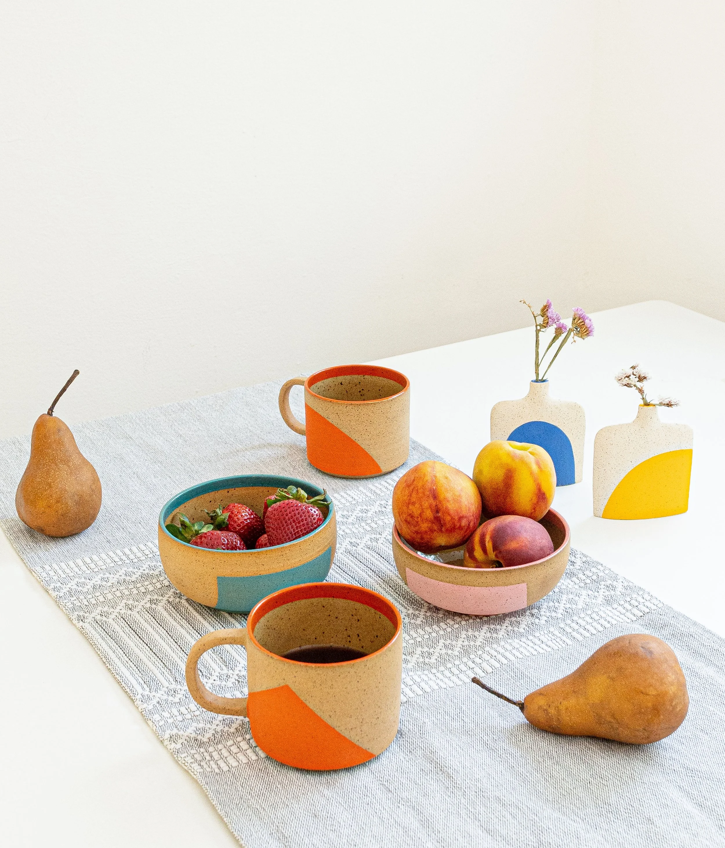 Tazas de cerámica con colores y formas geométricas, con frutas como duraznos y peras. Tazas con bebida, sobre un mantel de tela blanca con un fondo de pared blanca.