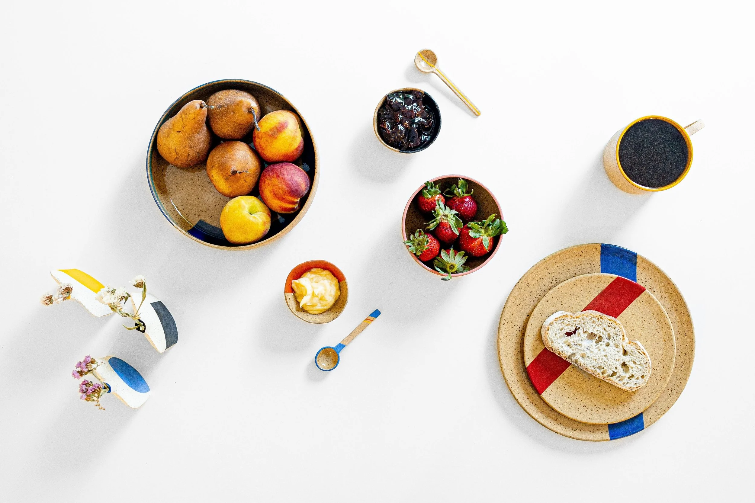 Mesa con frutas, mermelada, café, pan, mantequilla, y utensilios coloridos en platos y tazas de LODAZAL Cerámica