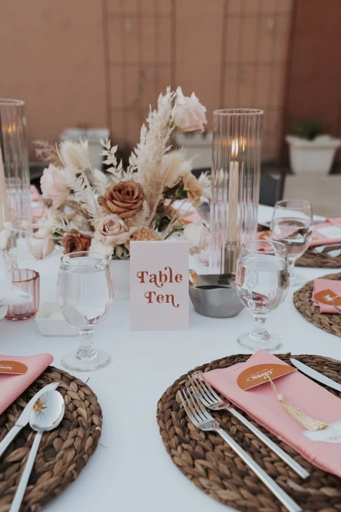 A table set for a celebration with a floral centerpiece, water glasses, woven placemats, pink napkins, and a sign reading 'Table Ten'.