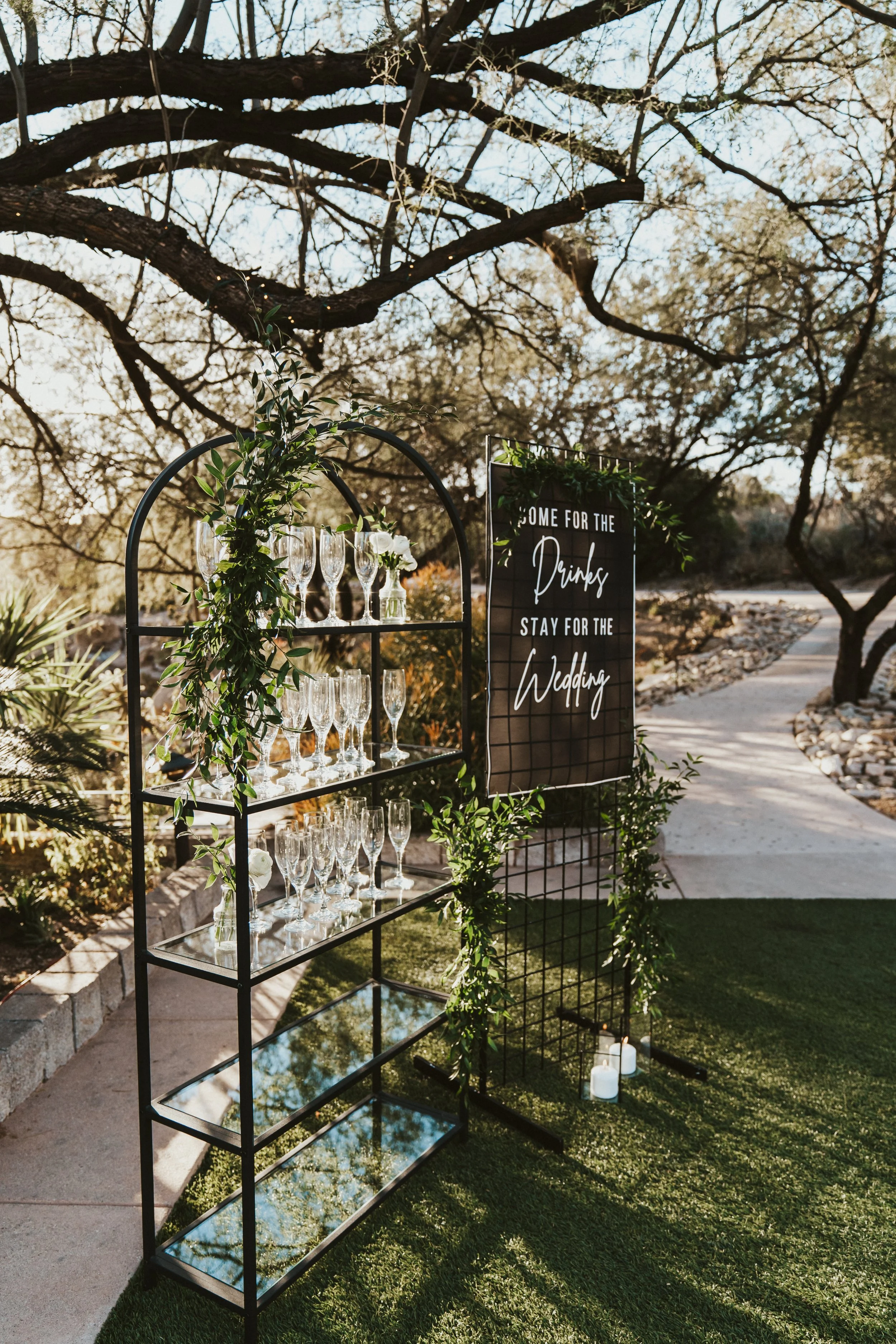 Outdoor wedding setup with glasses on a metal shelf and a decorative sign saying 'Come for the drinks, stay for the wedding.'