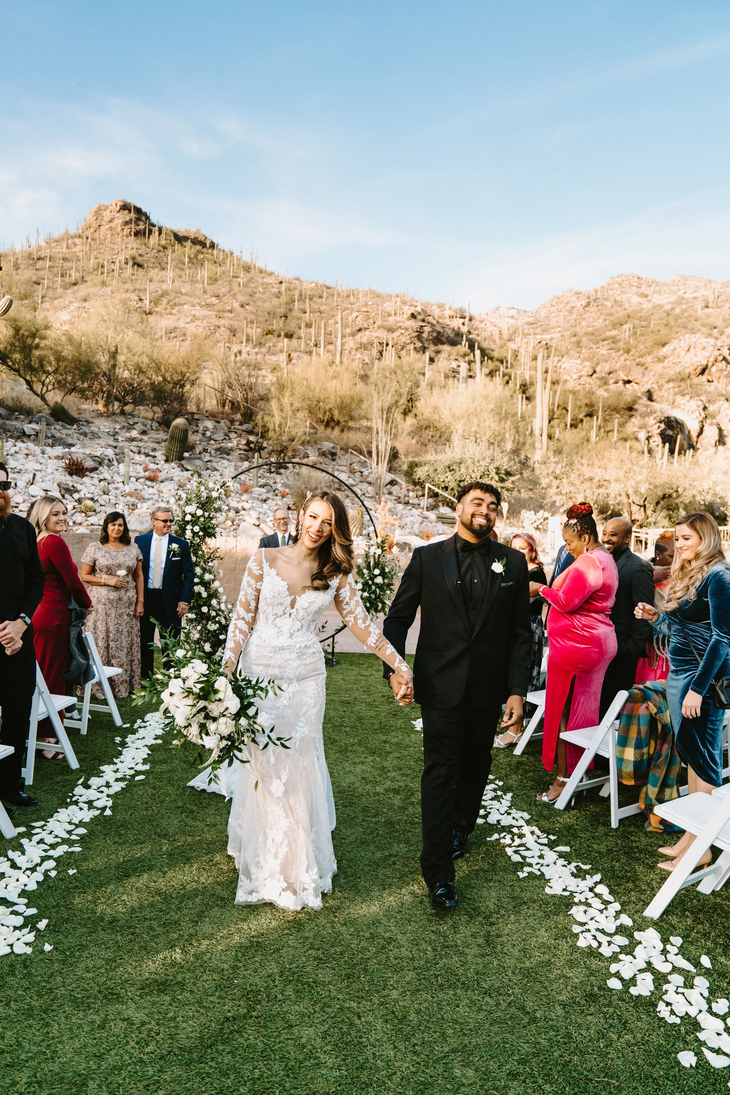 A bride and groom walking down an outdoor aisle, holding hands and smiling, surrounded by wedding guests. The wedding is set in a desert landscape with mountains and cacti in the background. The bride is wearing a long white dress with lace details, and the groom is in a black suit.