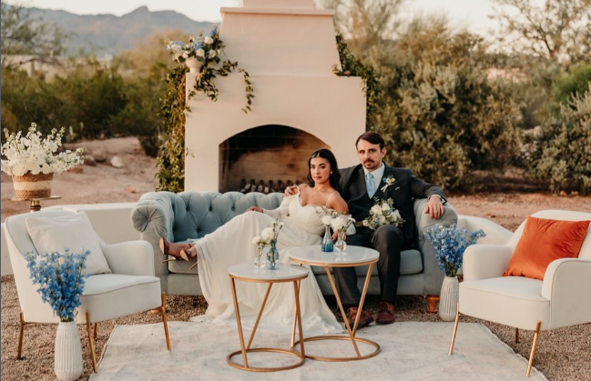 A bride and groom sitting on a gray vintage sofa outdoors near a white fireplace decorated with flowers, with additional white and blue flower arrangements and a white rug on the ground.