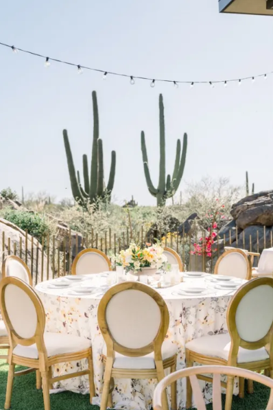 Outdoor dining area with a round table covered with a floral tablecloth, surrounded by cream-colored chairs, with desert plants and cacti in the background under a string of lights.