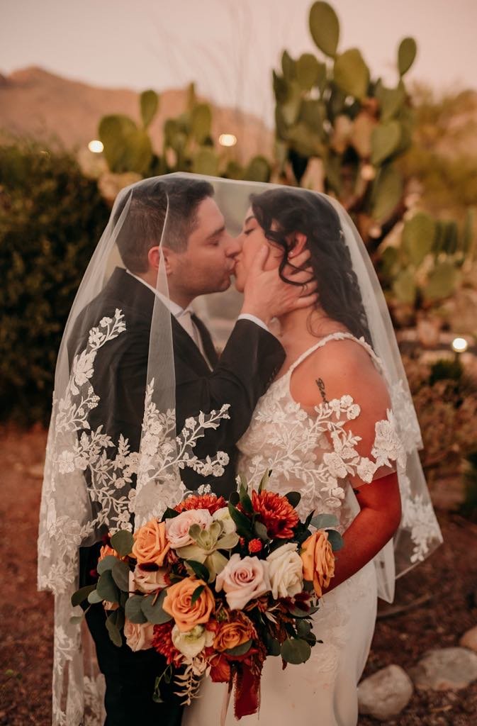 Bride and groom sharing a kiss under a lace veil, with the bride holding a bouquet of roses and other flowers, in a desert setting with cactus plants in the background.