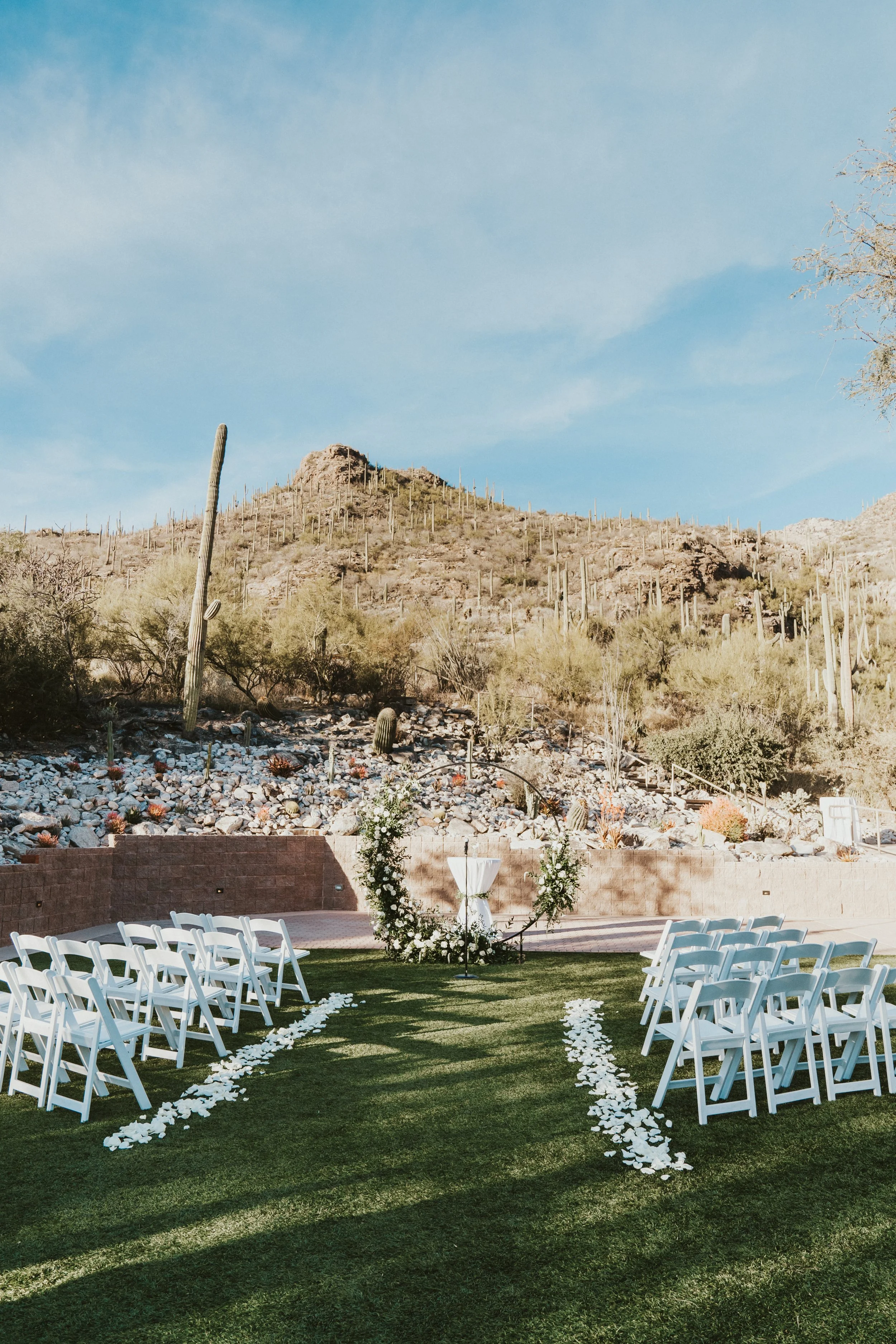 Outdoor wedding ceremony setup with white chairs and floral arch in a desert landscape, featuring cacti and rocky terrain under a blue sky.