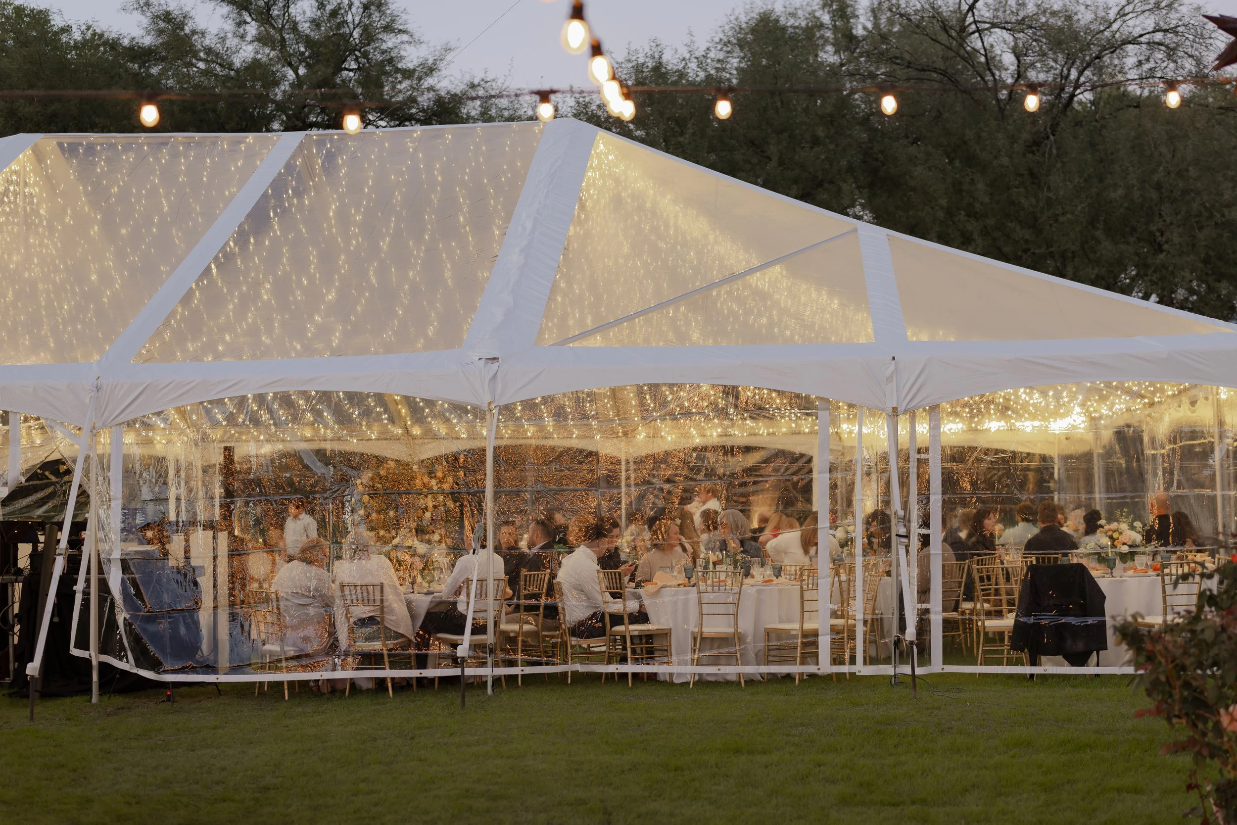 People dining at a wedding reception inside a large white tent decorated with string lights, with trees in the background.