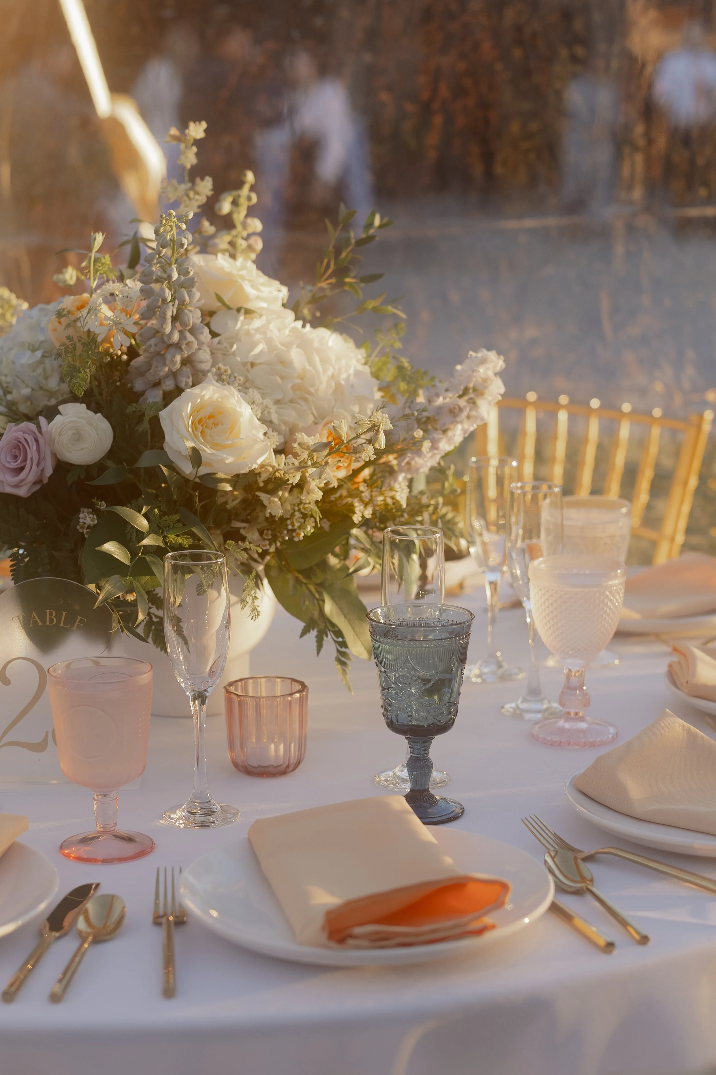 Elegant table setting with a large floral centerpiece, champagne glasses, pink and clear goblets, a salmon-colored cloth napkin on a white plate, and gold cutlery, set outdoors with a lake and trees in the background.