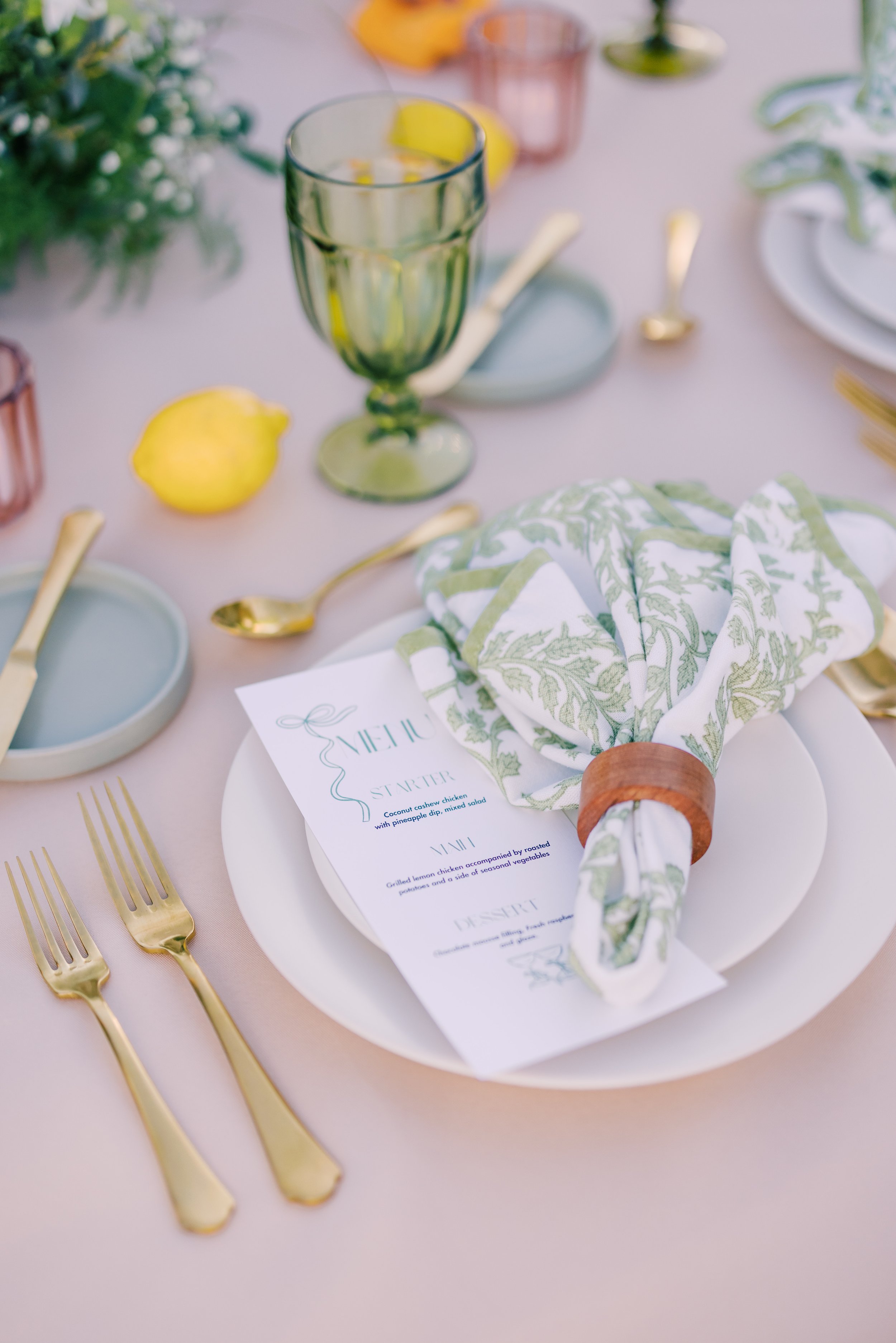 Elegant table setting with gold utensils, a green glass, a white plate with a green and white napkin, and a printed menu on a pink tablecloth, with a lemon and a flower arrangement in the background.