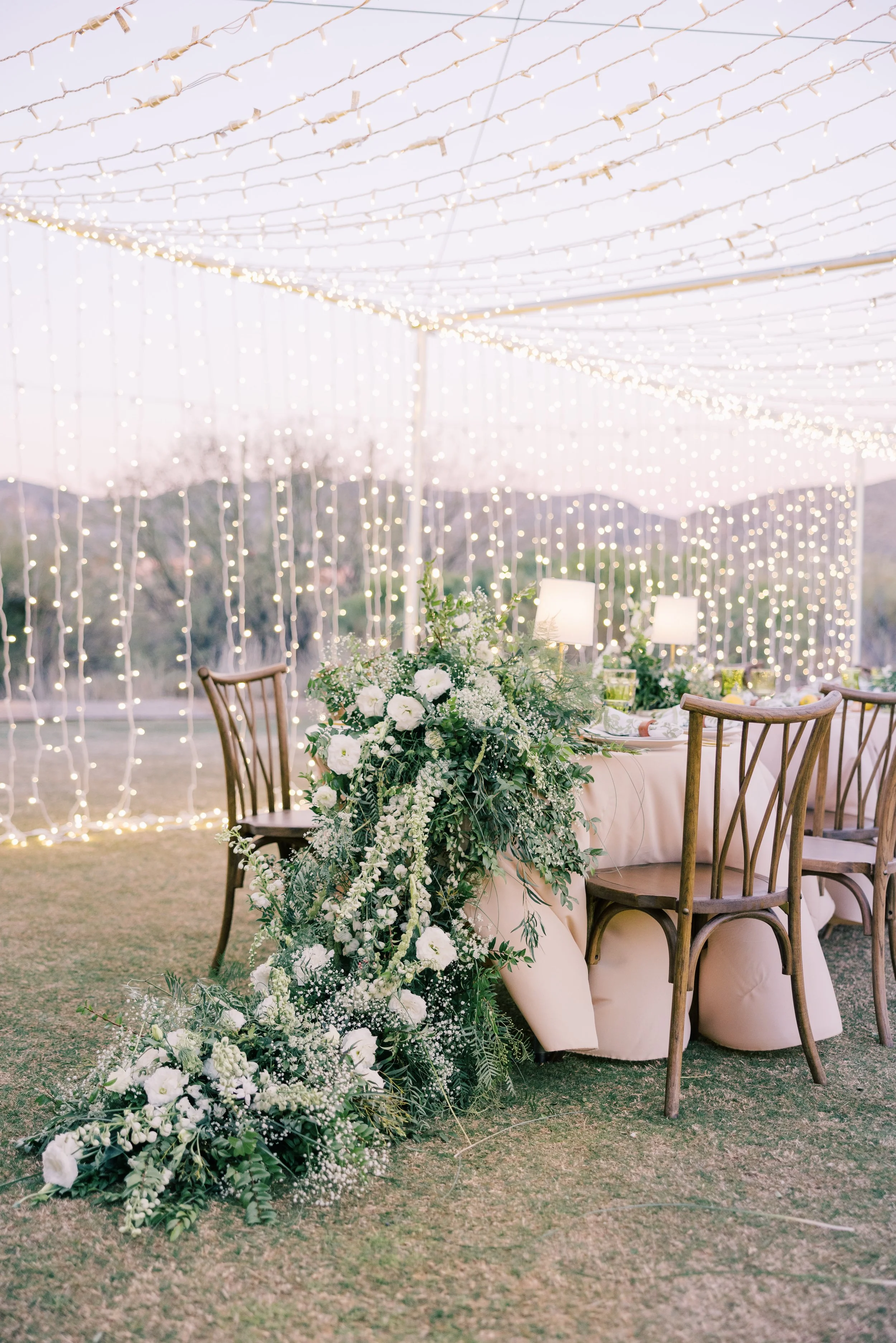 Outdoor wedding reception setup with a table decorated with a white tablecloth, surrounded by wooden chairs, under a canopy of string lights, and adorned with a large floral arrangement of white flowers and greenery.
