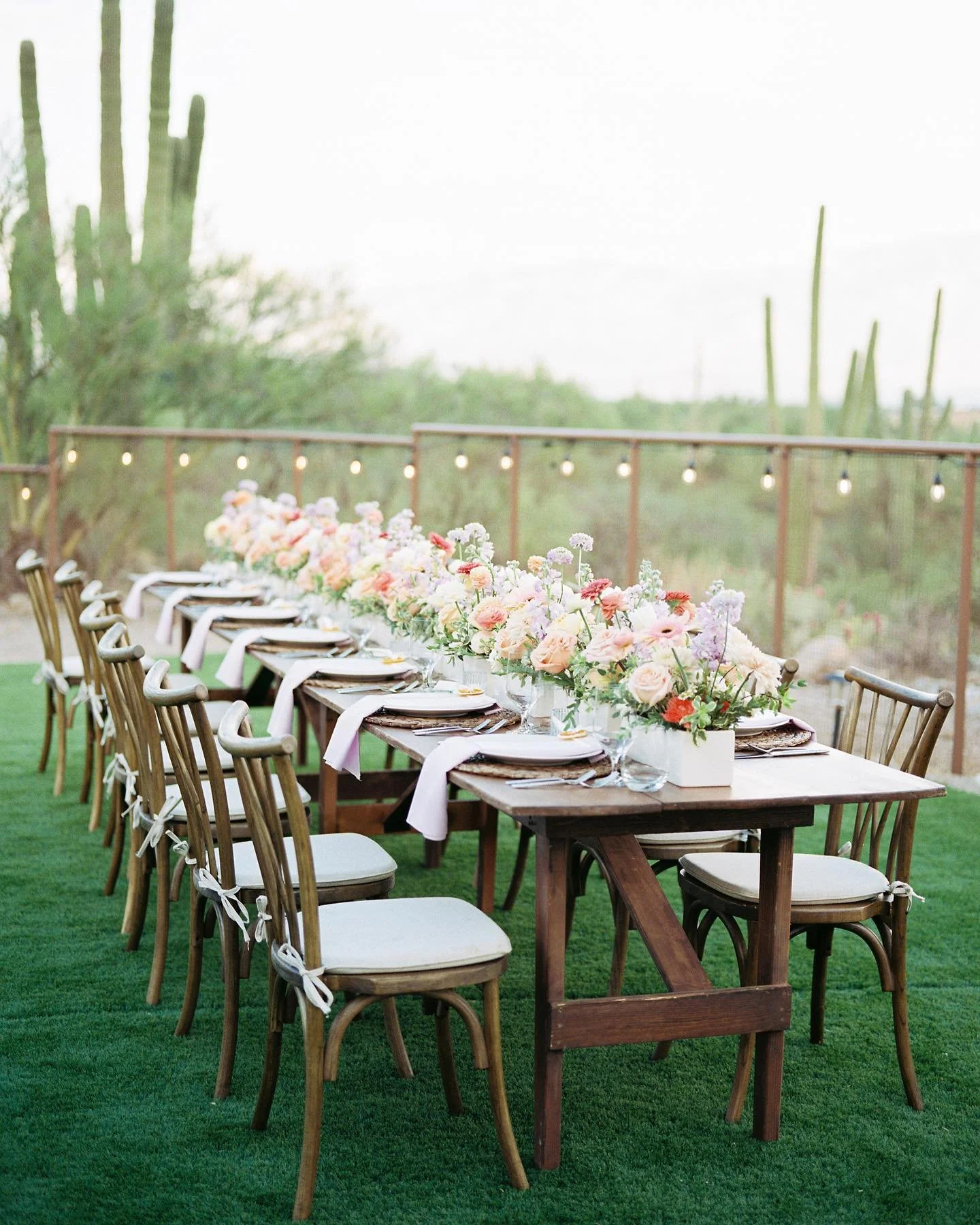 Outdoor event table decorated with floral arrangements, white napkins, and tableware, set on a grassy area with string lights and cactuses in the background.