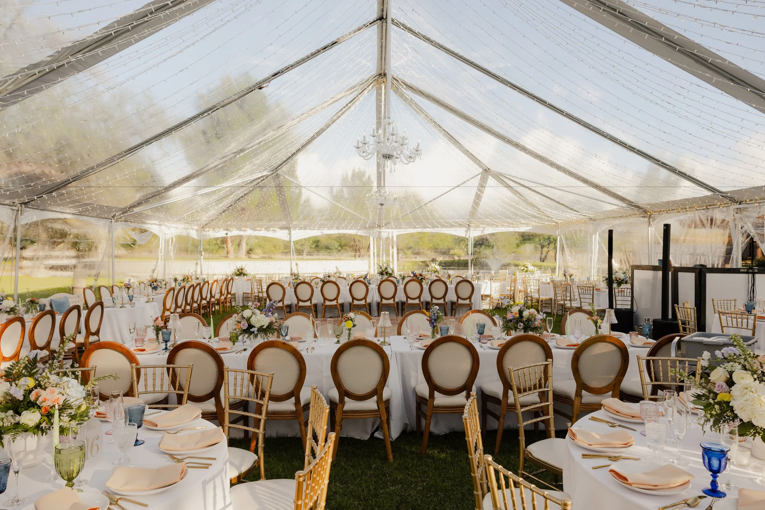 Elegant event setup inside a large transparent tent with round and rectangular tables, decorated with floral arrangements, tableware, and chairs, prepared for a formal gathering or wedding reception.