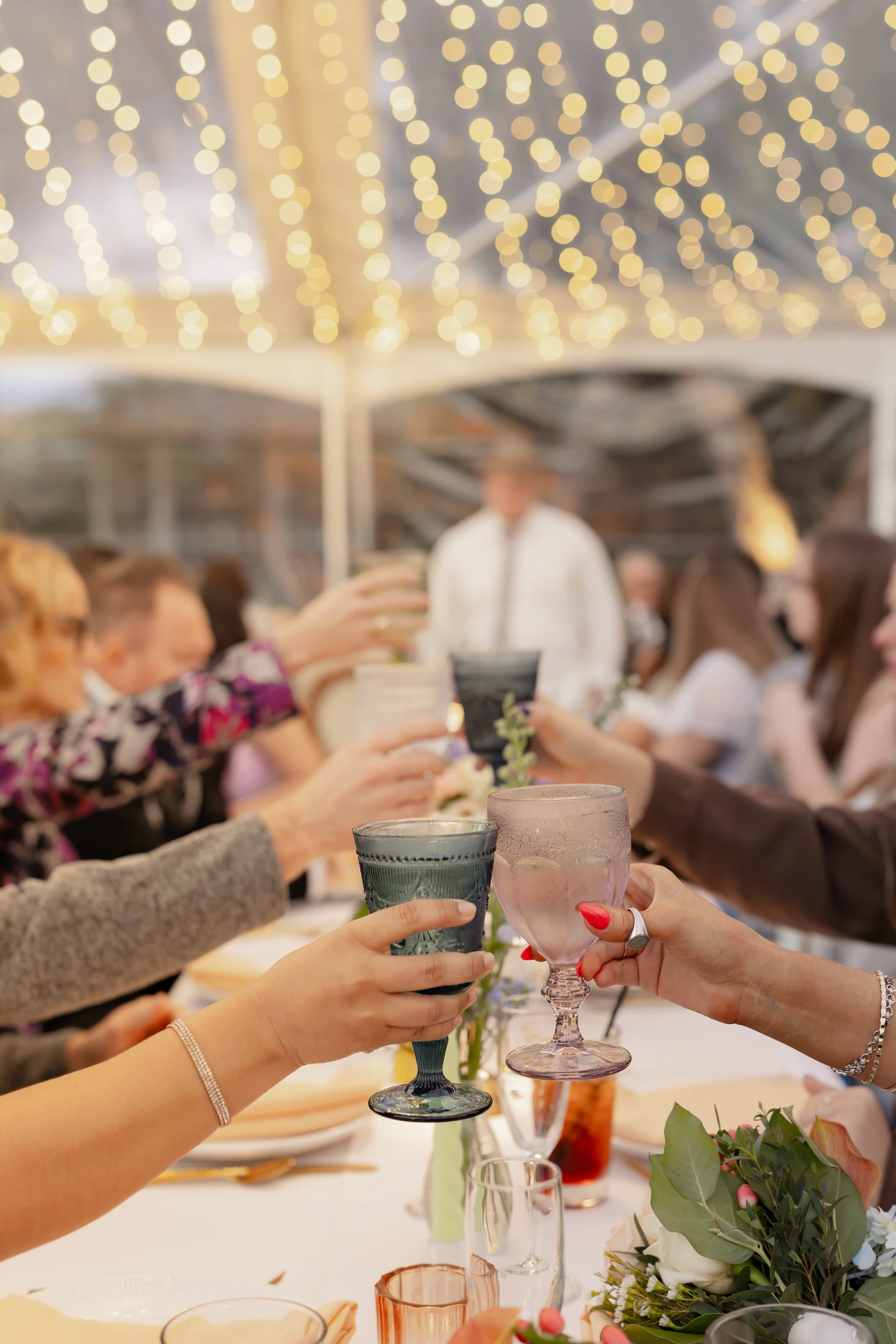 People raising glasses in a toast at a celebration dinner under string lights in a decorated outdoor setting.