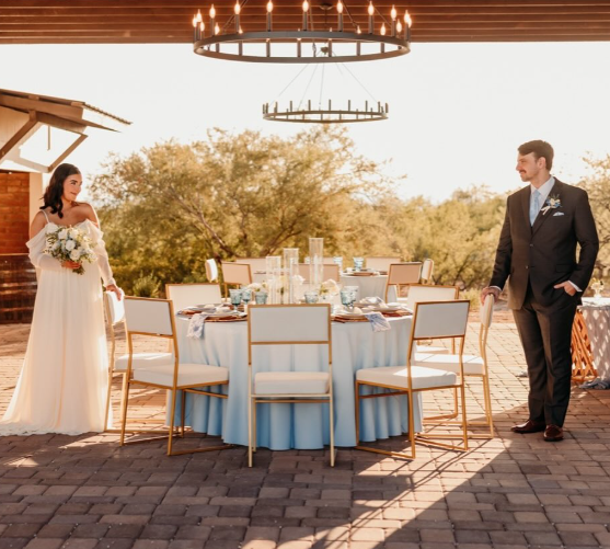 Bride and groom standing across a round, elegantly set table at an outdoor wedding reception with trees in the background