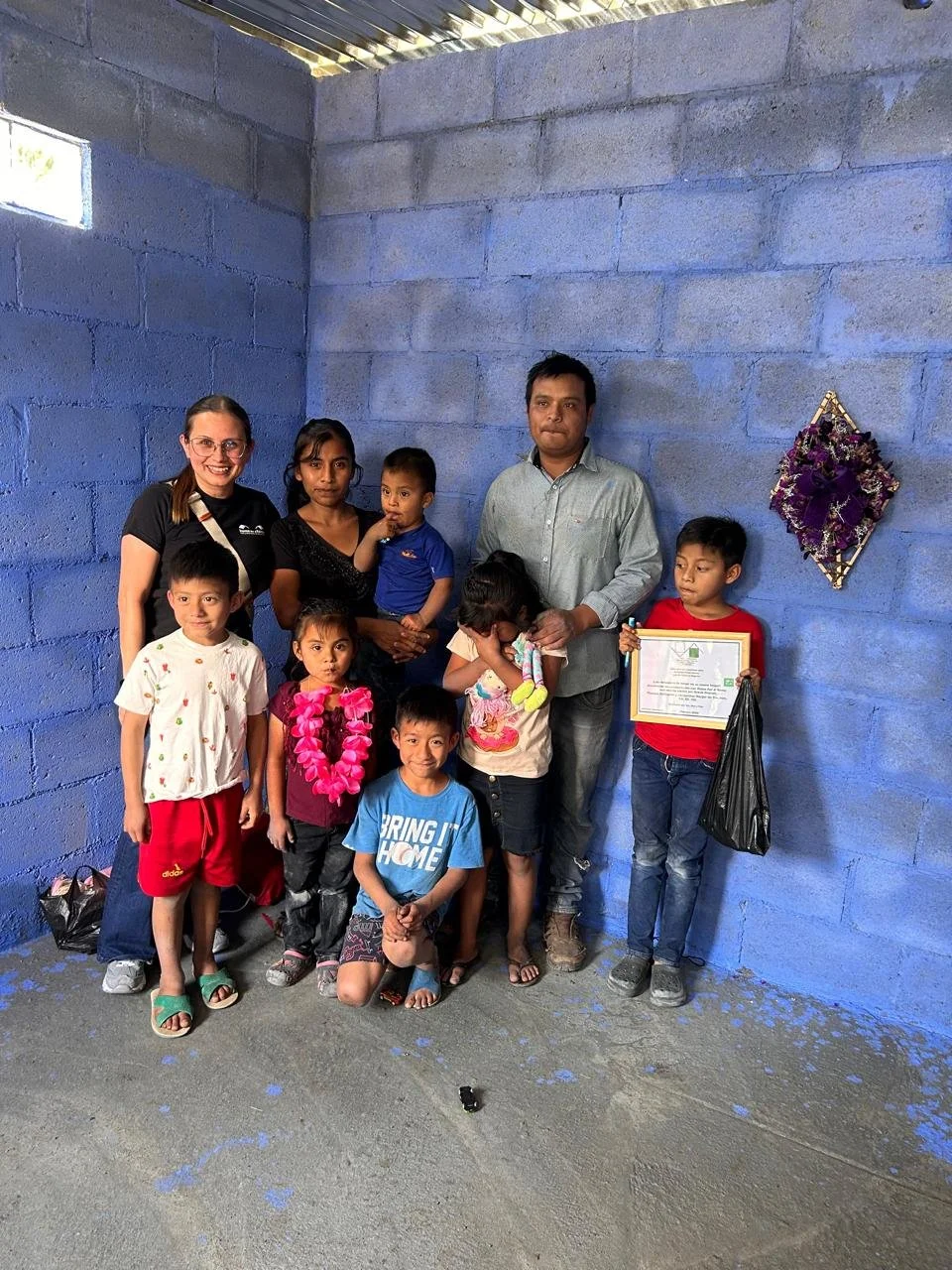 A group of ten people, including children and adults, standing together against a blue brick wall. One child holds a framed certificate, and a woman is wearing a pink flower lei. There are purple floral decorations on the wall.