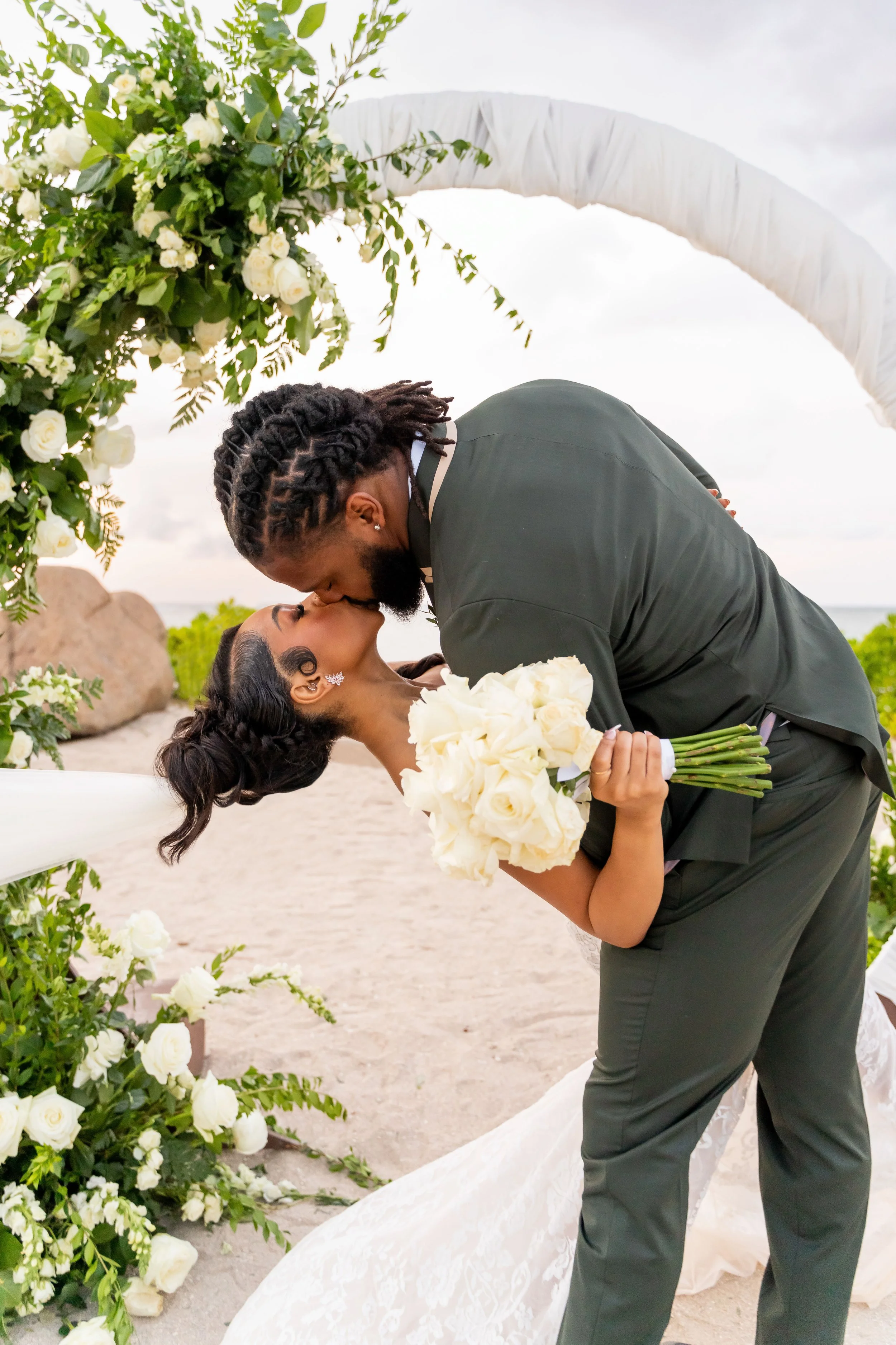 A couple sharing a kiss at a wedding ceremony on the beach, with floral decorations and an arch.