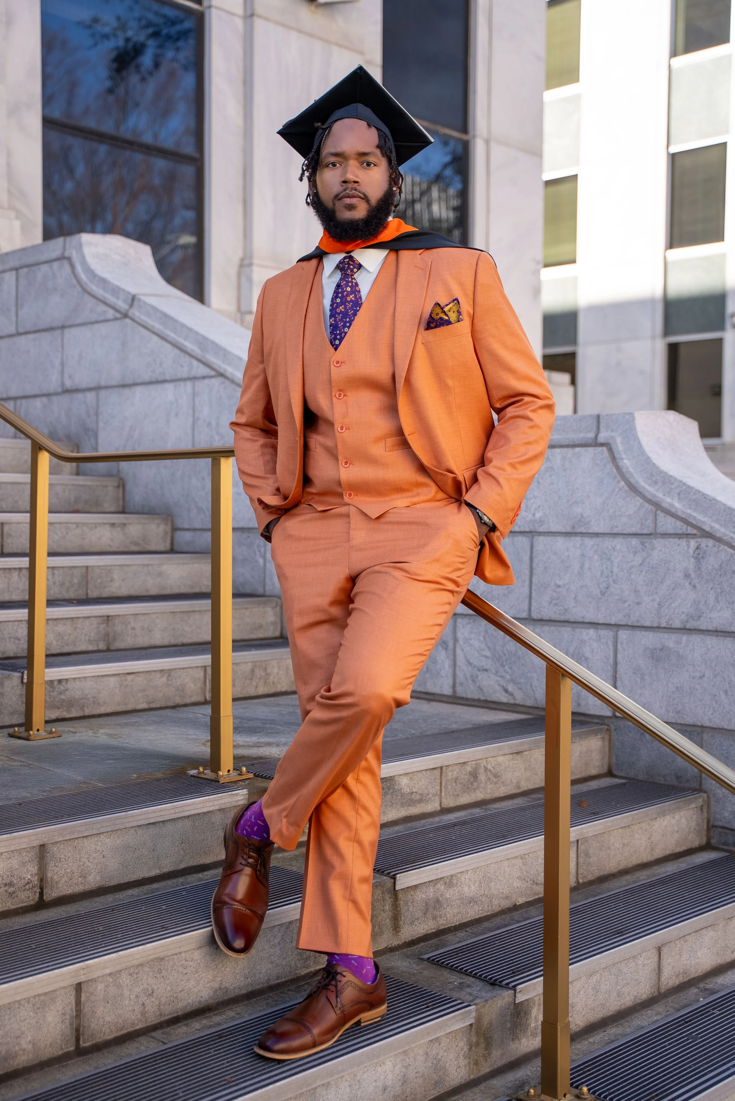 A man dressed in an orange suit with a patterned tie, pocket square, and dress shoes, standing on stairs outside a building, wearing a graduation cap and gown.