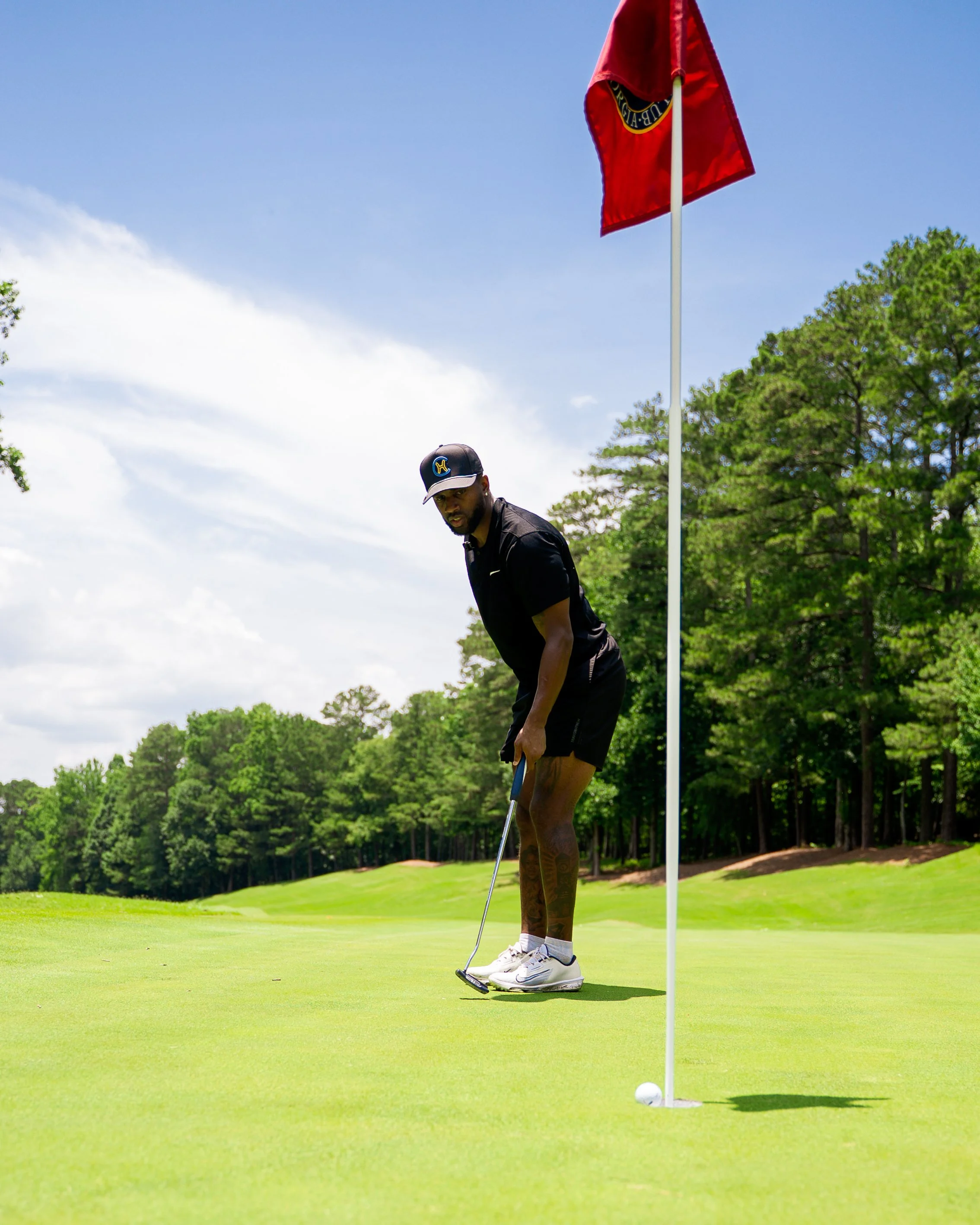 A man preparing to putt on a golf course near a flagstick with a golf ball nearby, surrounded by trees and a partly cloudy sky.