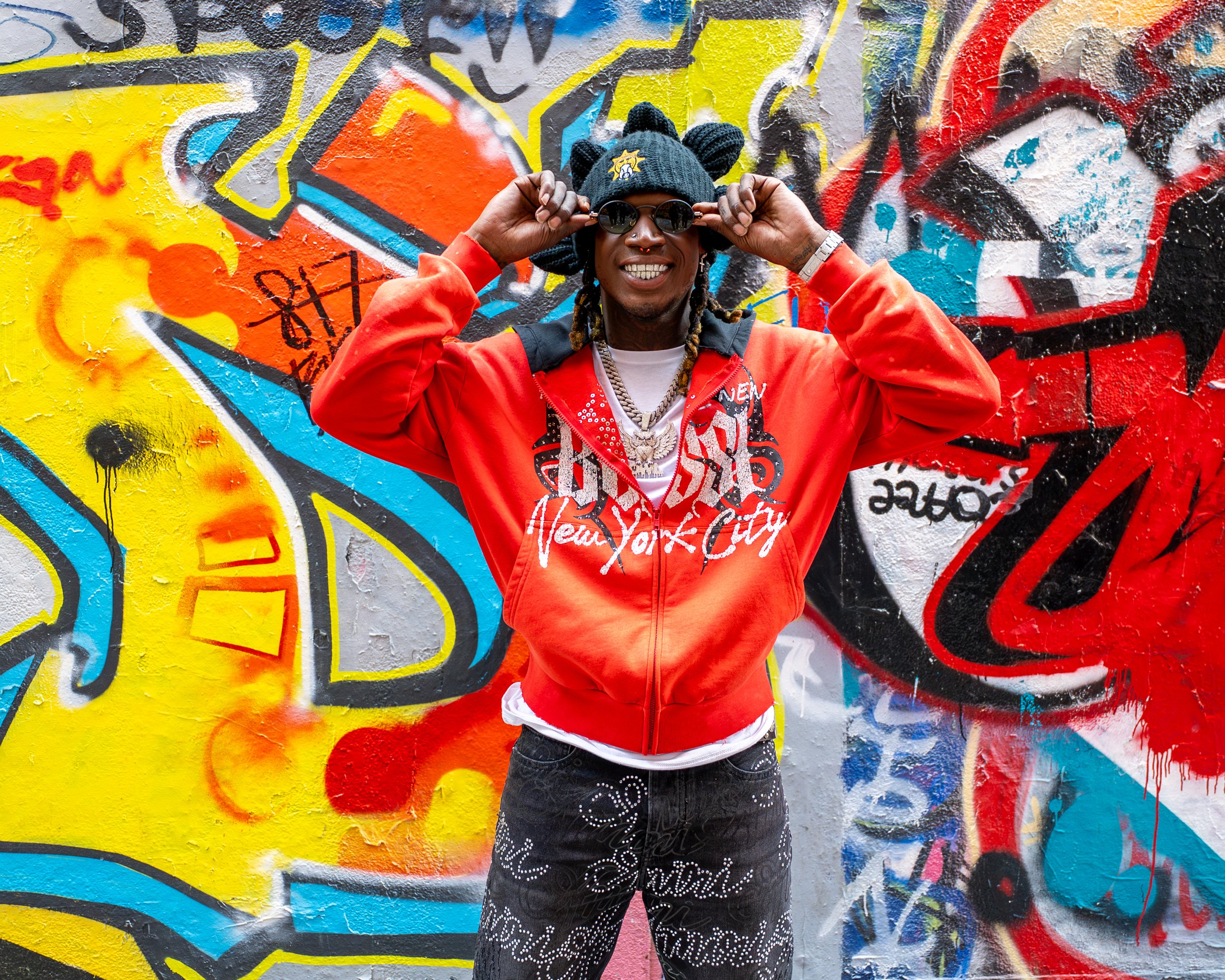 A man standing in front of a colorful graffiti wall. He is wearing a black beanie with a badge, sunglasses, a red hoodie with 'New York City' written on it, and patterned black jeans. He is smiling and holding his sunglasses with both hands.