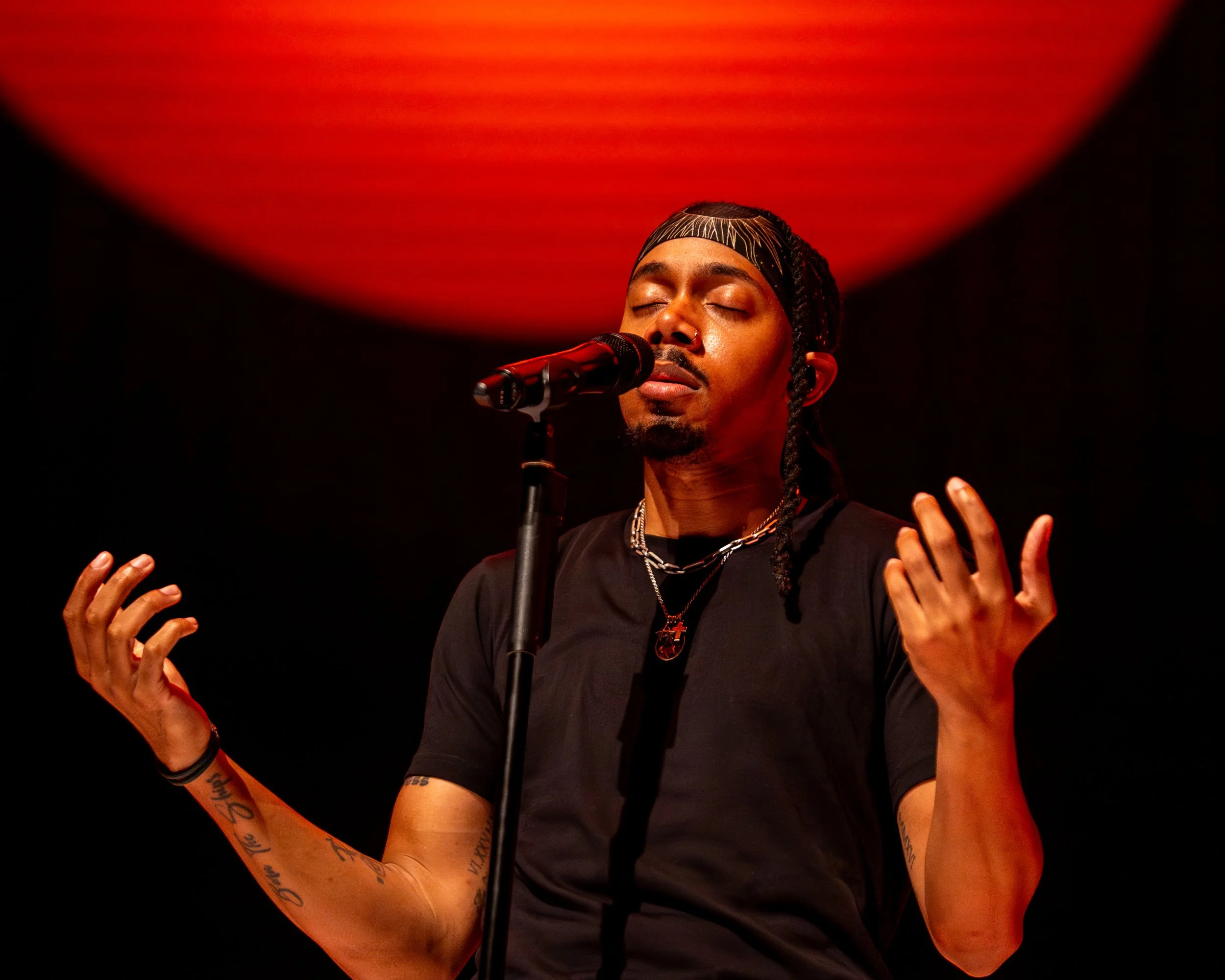 A man with dreadlocks and tattoos on his arms performs on stage with eyes closed, singing into a microphone, with a red circle light behind him.