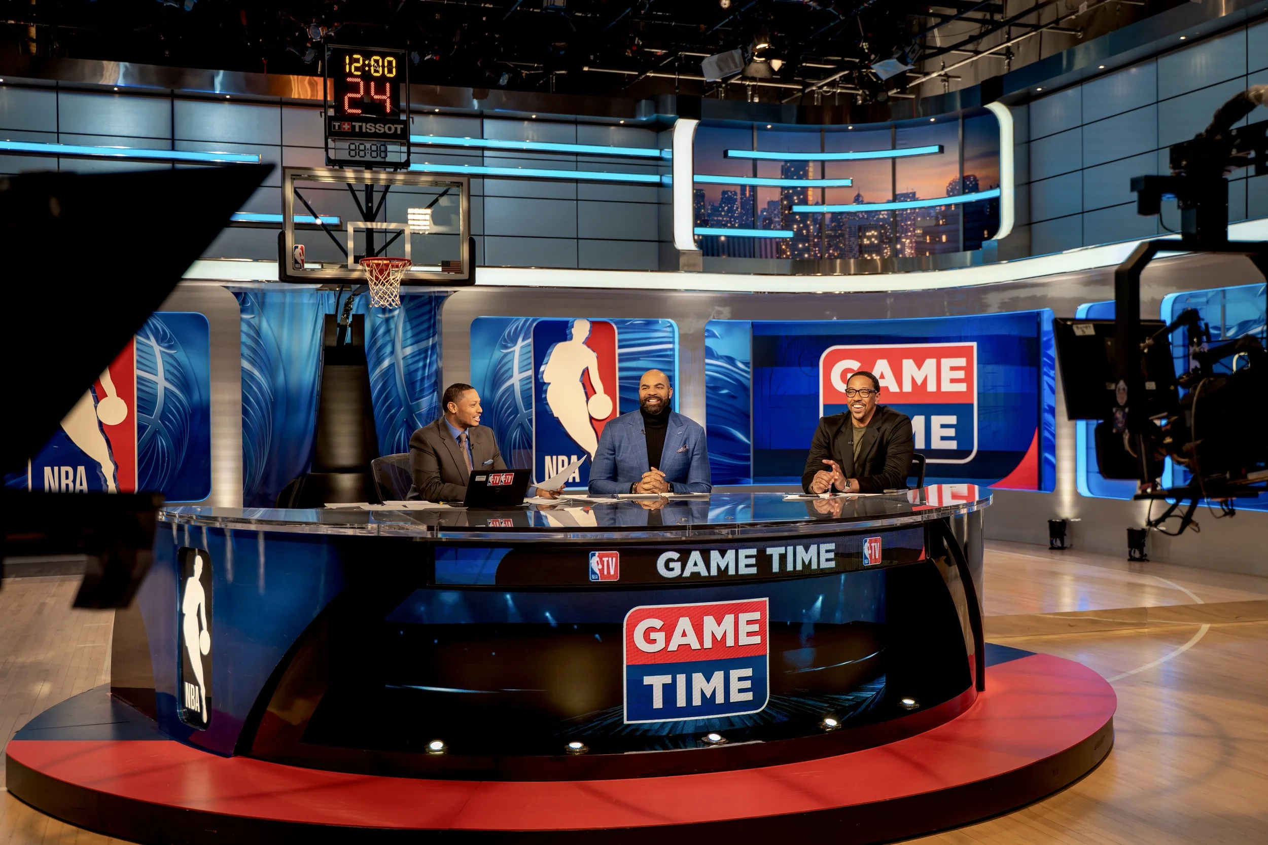 Three men sitting at a curved desk with NBA, NBA TV, and Game Time logos, on a TV set with basketball court and city skyline backdrop, studio lighting, and television cameras.