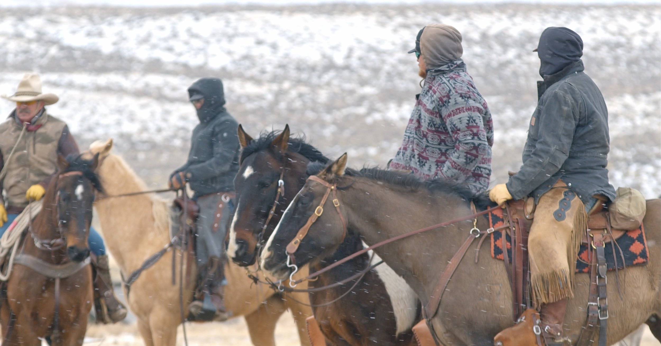 Group of cowboys riding horses in snowy landscape, wearing winter clothing.
