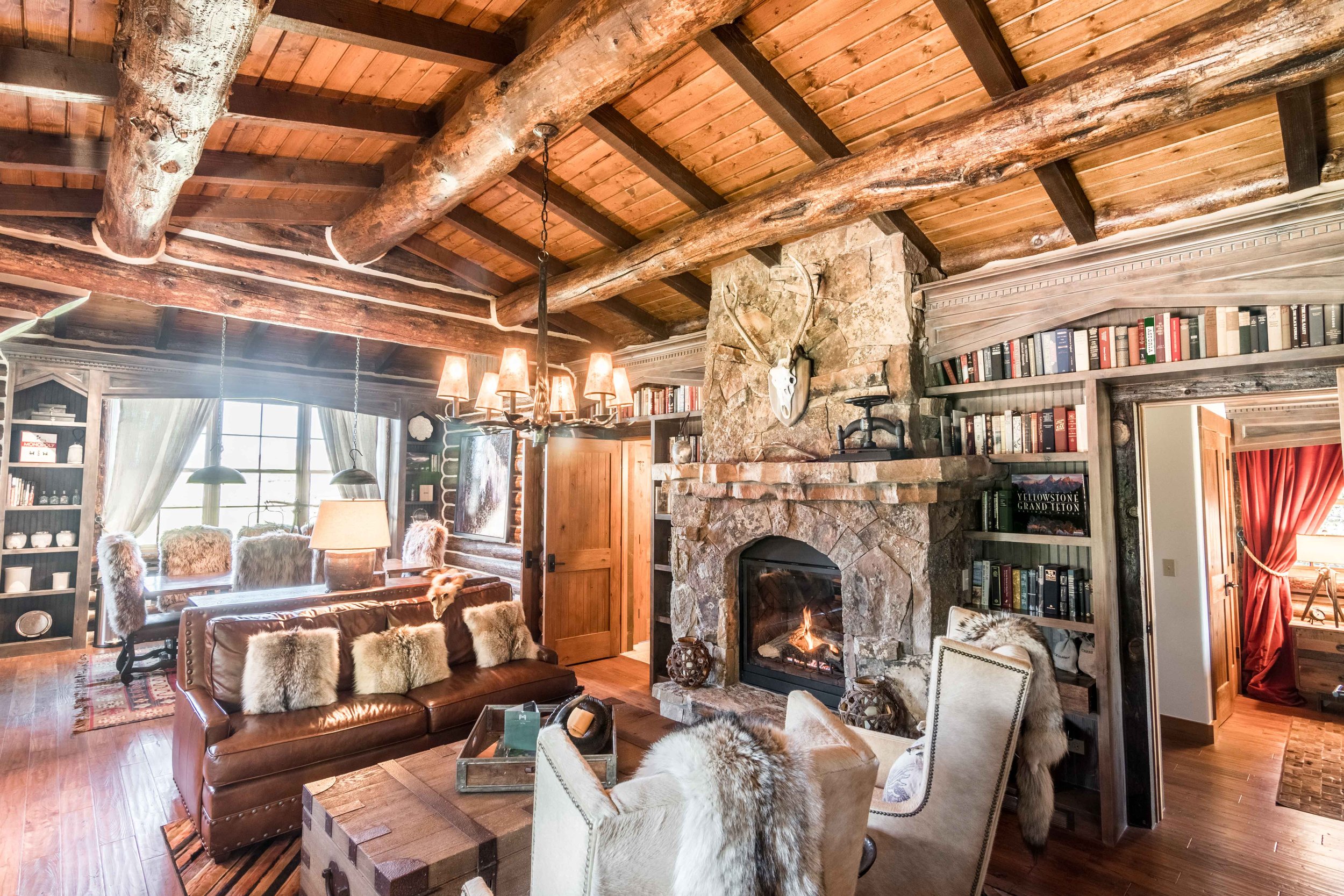 Rustic cabin interior with stone fireplace, wooden beams, leather couch with fur cushions, log ceiling, chandelier, bookshelf, and large window.