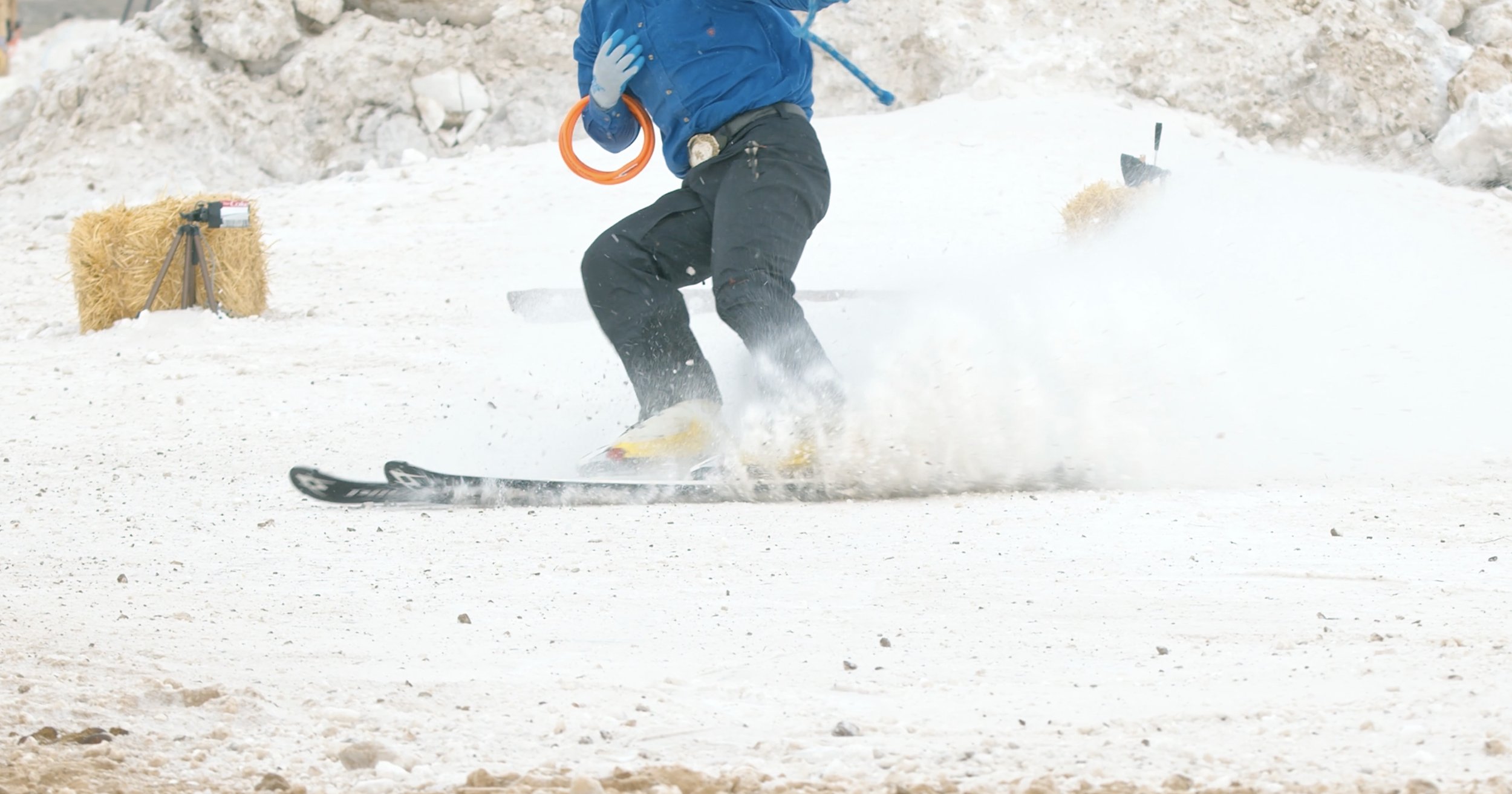 Person skiing on snow with a stick and an orange ring in hand, creating snow splashes, hay bales and tripod in background.