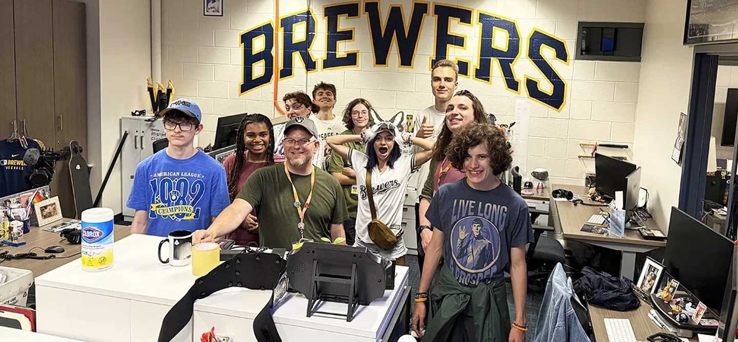 IOB students and mentors visit the Milwaukee Brewers office during CollabQuest, posing together beneath a large Brewers wall sign.