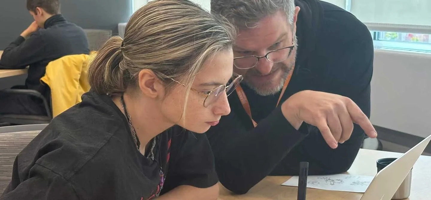An IOB mentor works one-on-one with a student during Quest, reviewing creative work on a laptop and offering guidance on an advanced digital project.