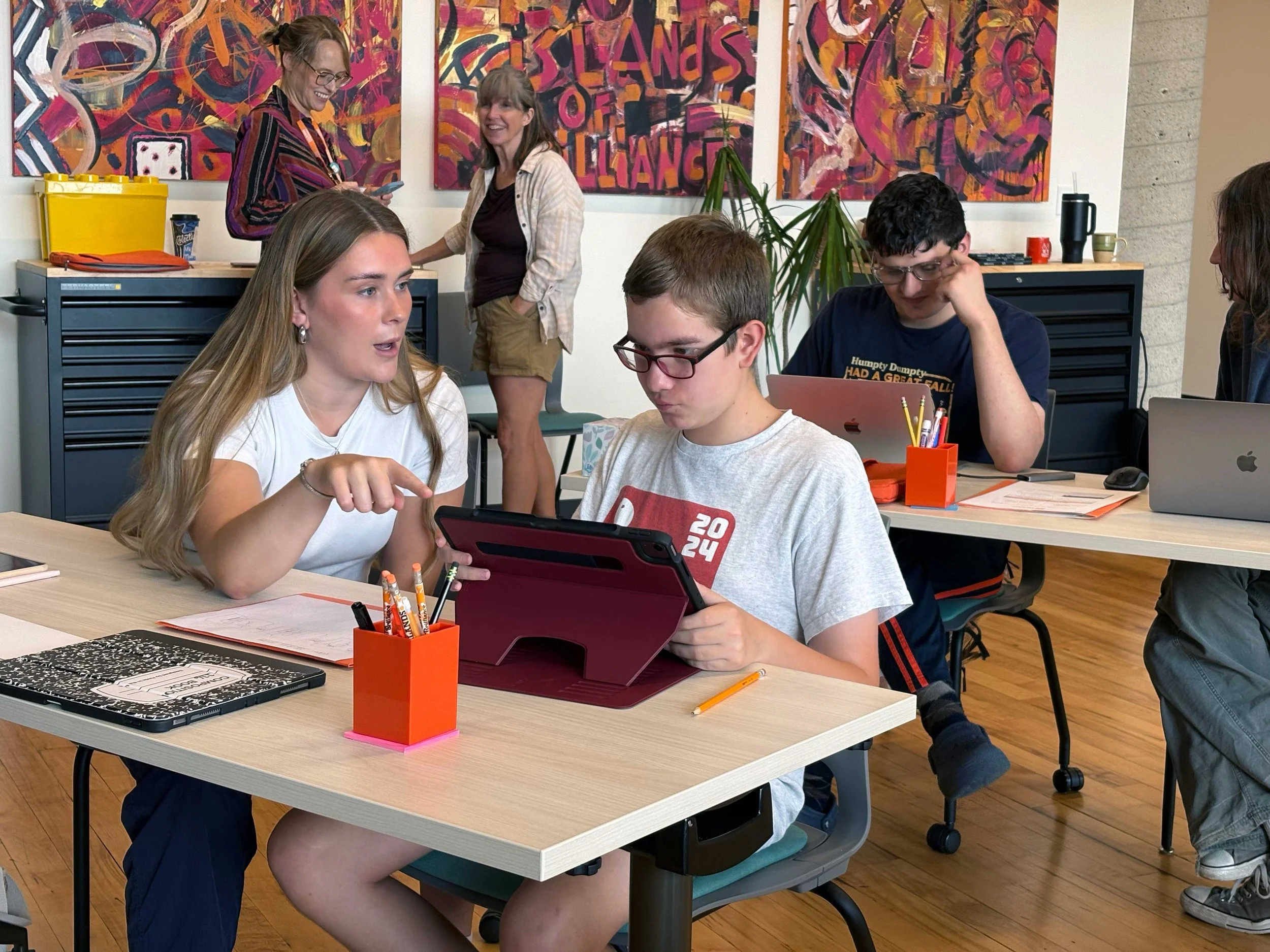 Students and mentors work together in an IOB Foundation workshop, collaborating on iPads and laptops in a colorful studio with artwork on the walls.