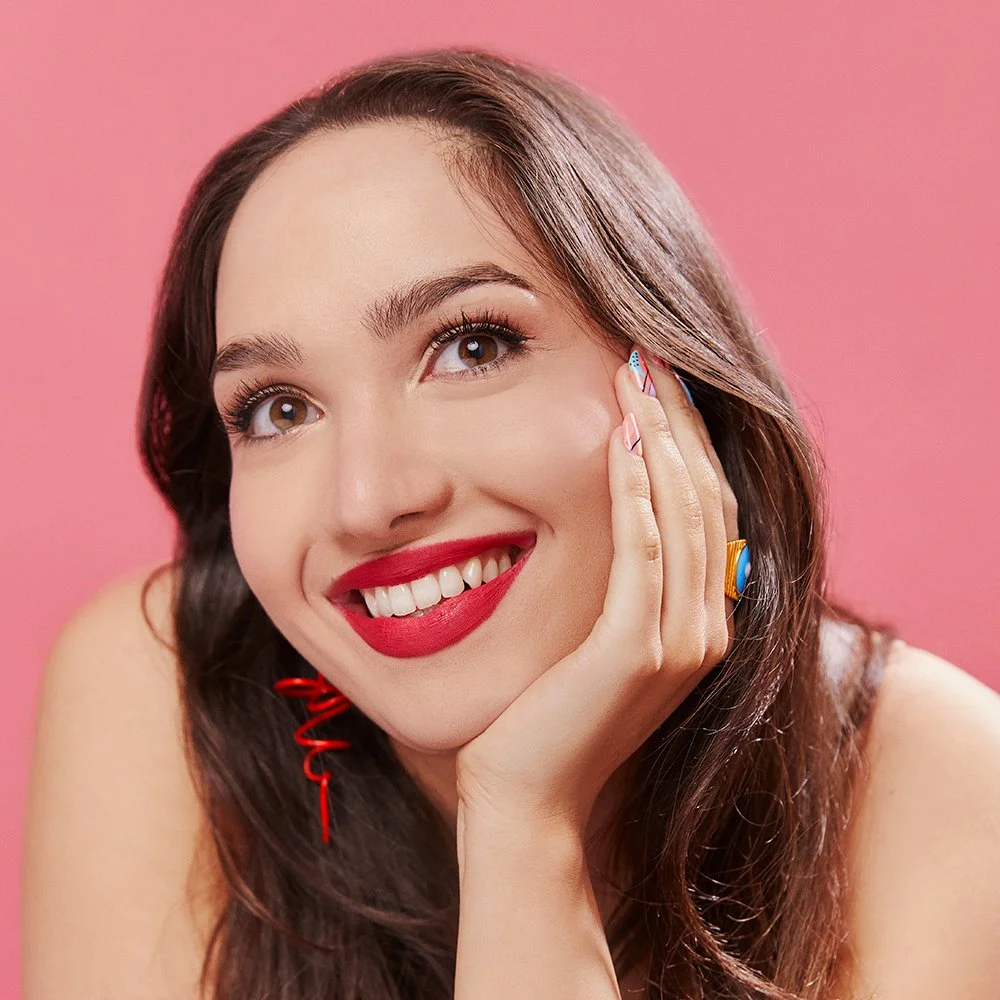 Close-up of a smiling woman with long brown hair, red lipstick, and colorful earrings against a pink background.