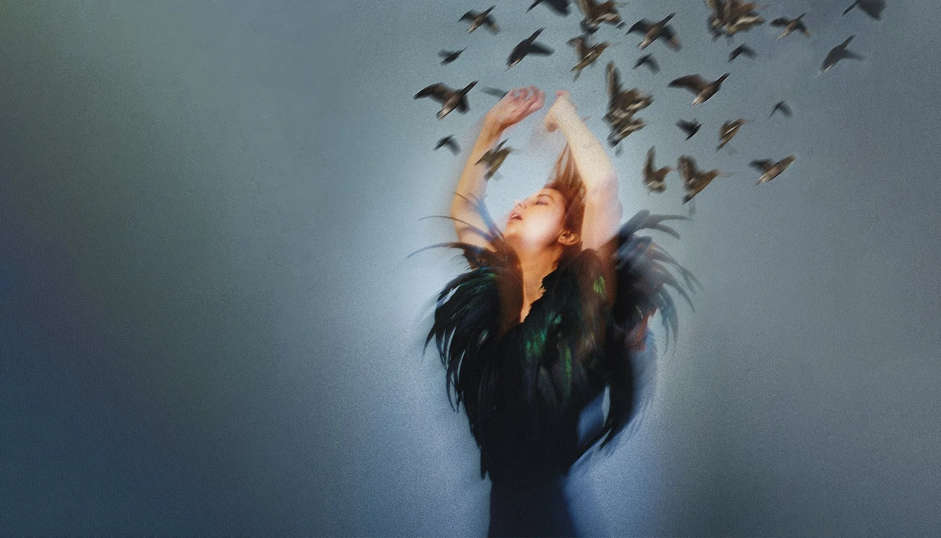 A woman with long brown hair, dressed in a black feathered outfit, has her arms raised above her head and eyes closed as a flock of small birds fly around her against a light gray background.