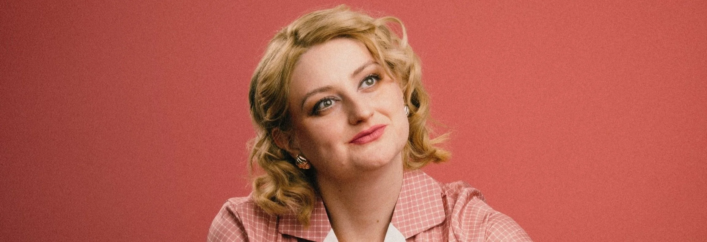 A woman with blonde curly hair and blue eyes, a pink checkered shirt, and earrings, sitting against a solid pink background.