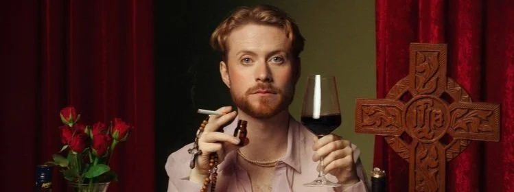 A man with a beard holding a glass of red wine and a cigarette, sitting at a table with red roses, a bottle, and a large wooden cross in the background.